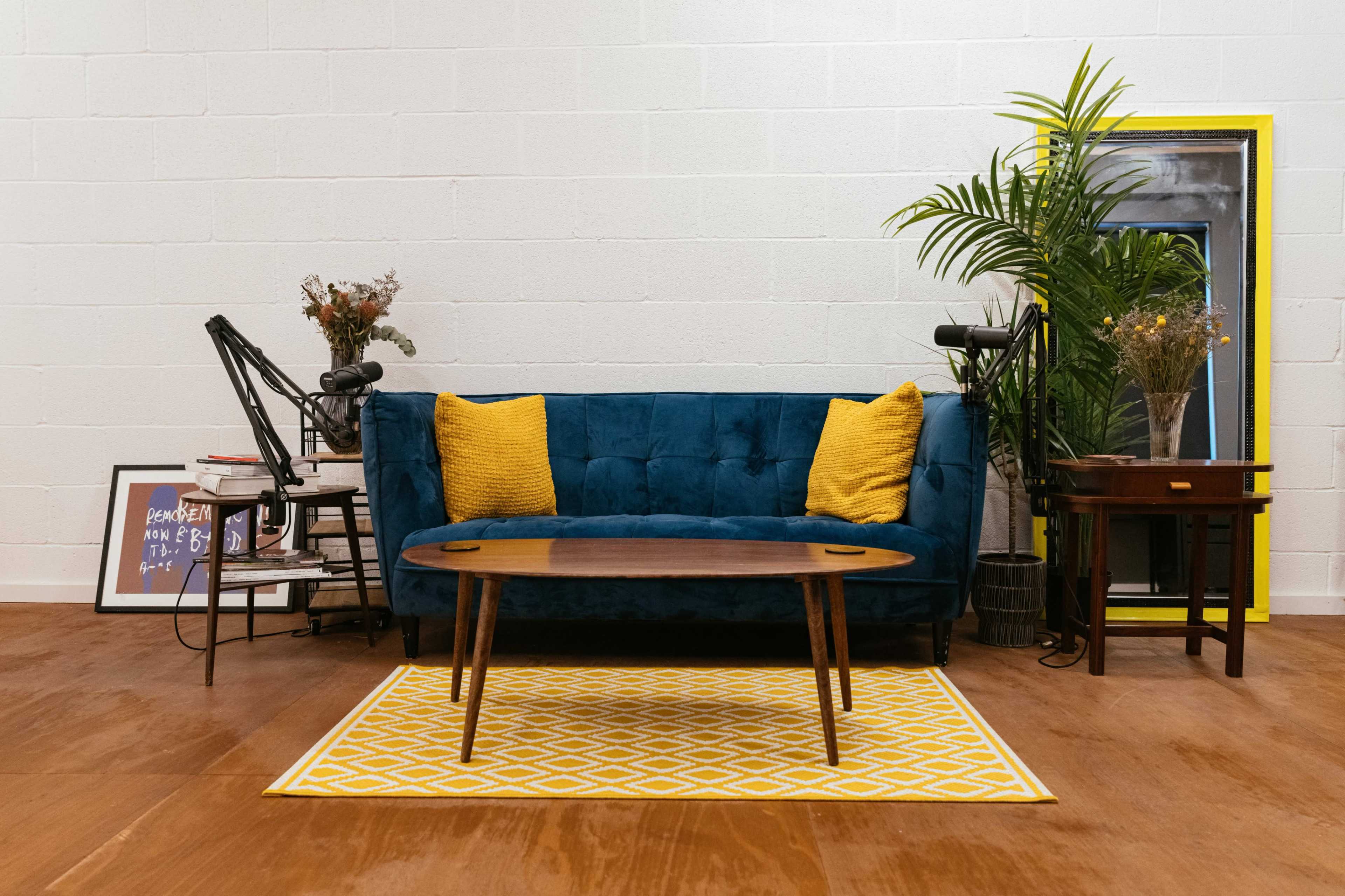 A blue sofa with yellow cushions sits in a living room, accompanied by a wooden coffee table, decorative plants, and framed artwork.