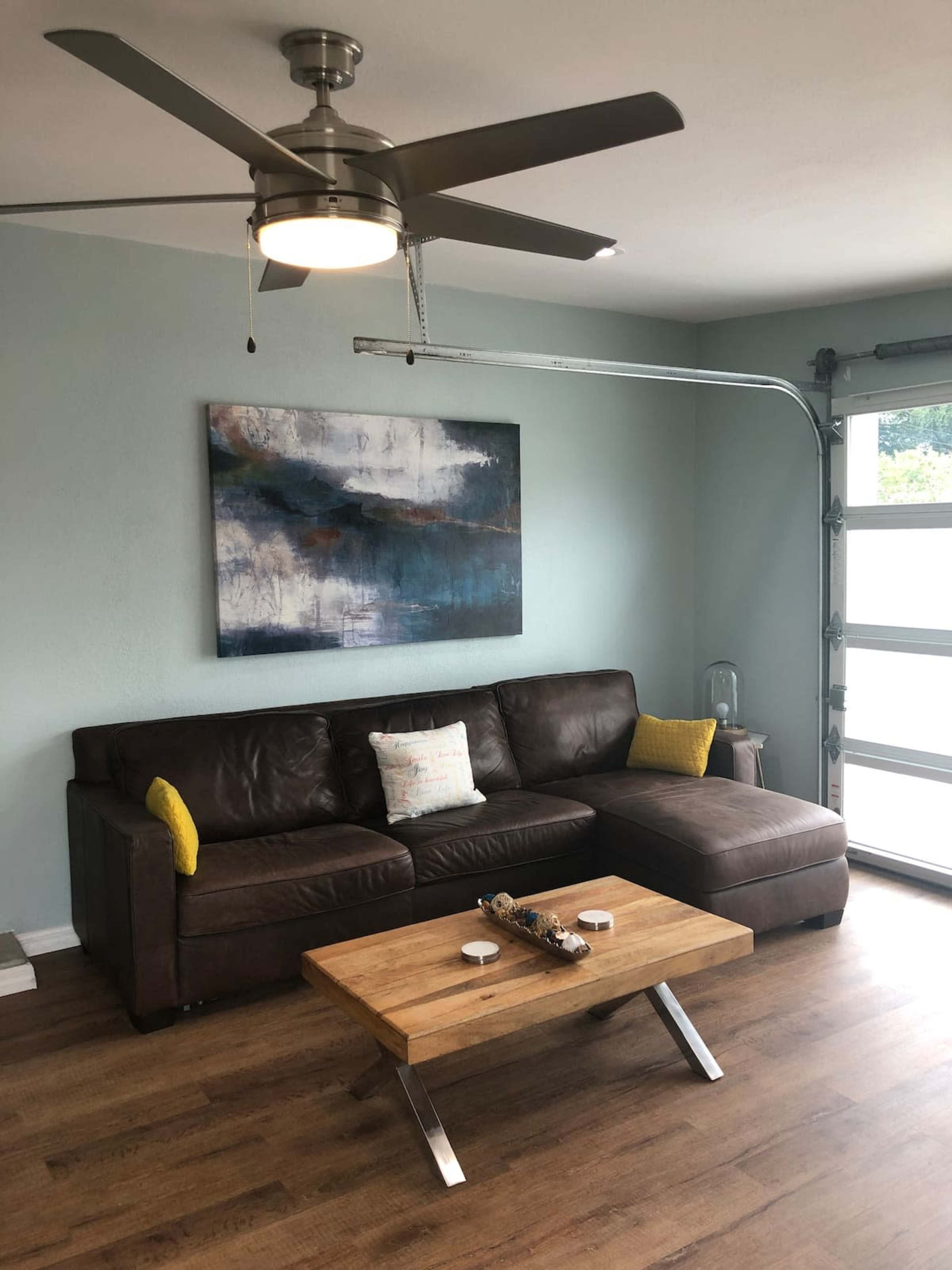 A living room features a brown leather sectional couch, a wooden coffee table, and a modern ceiling fan, with a large abstract painting on the wall.