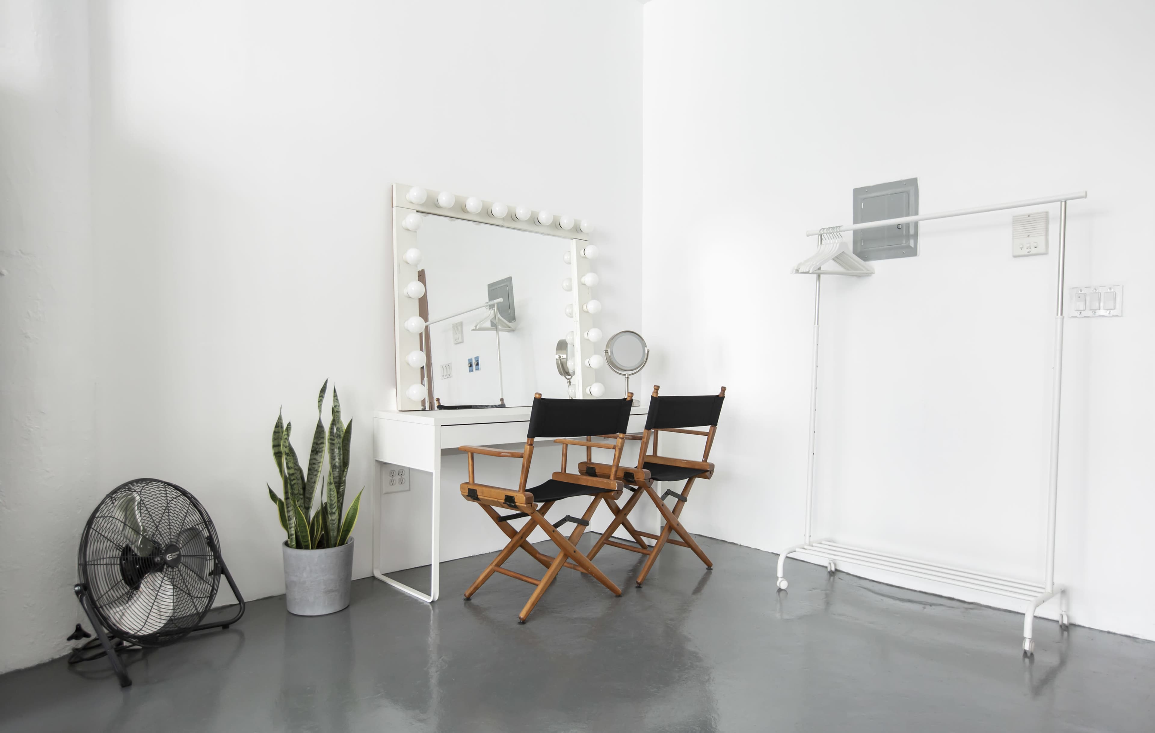 The image shows a minimalistic makeup vanity area with two director's chairs, a mirror framed with bulbs, a floor fan, a potted plant, and a clothes rack against a white wall.