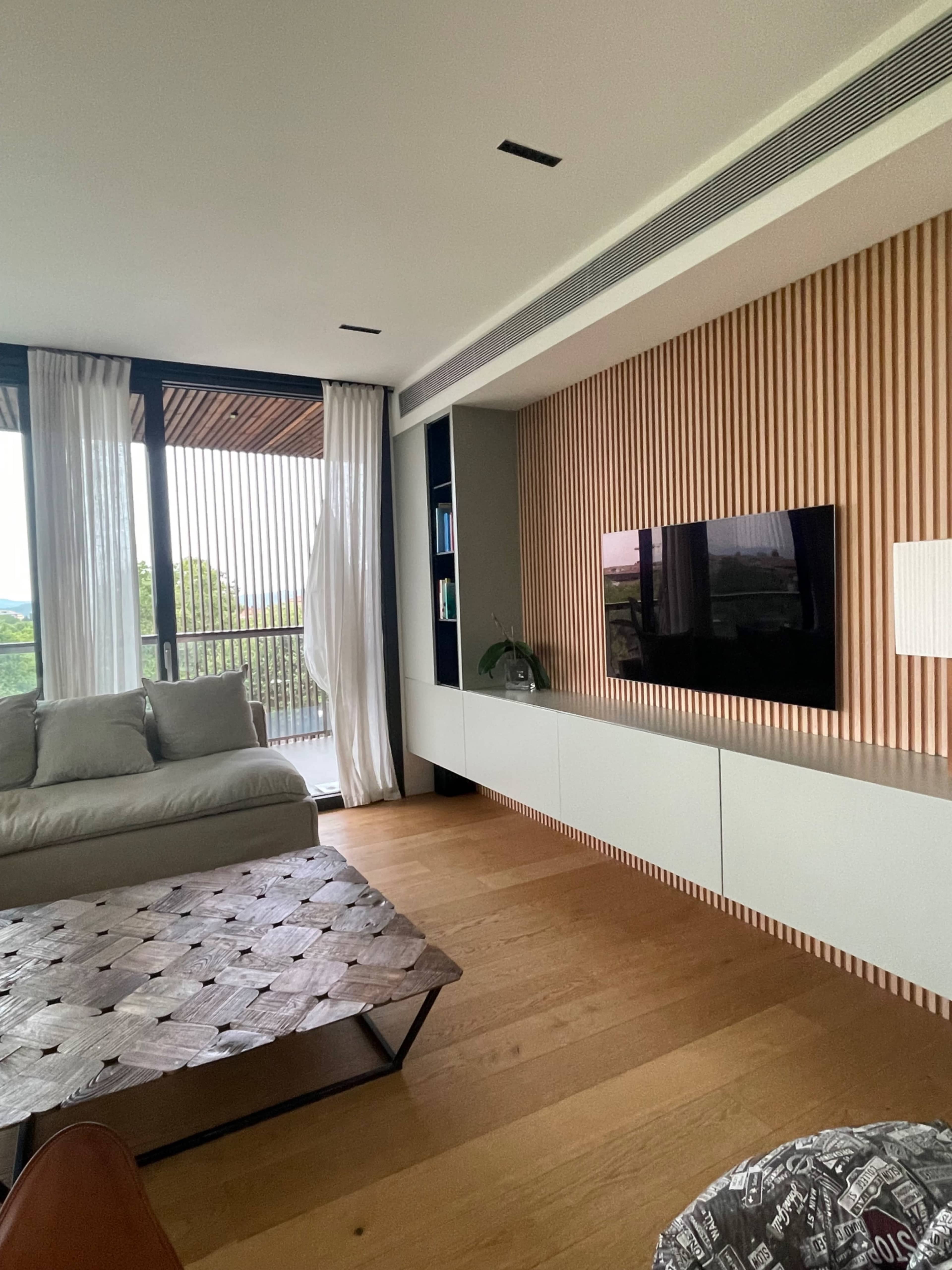 The image shows a modern living room featuring a light-colored sofa, a coffee table with a leather top, and a wall-mounted television against a textured wood panel backdrop.