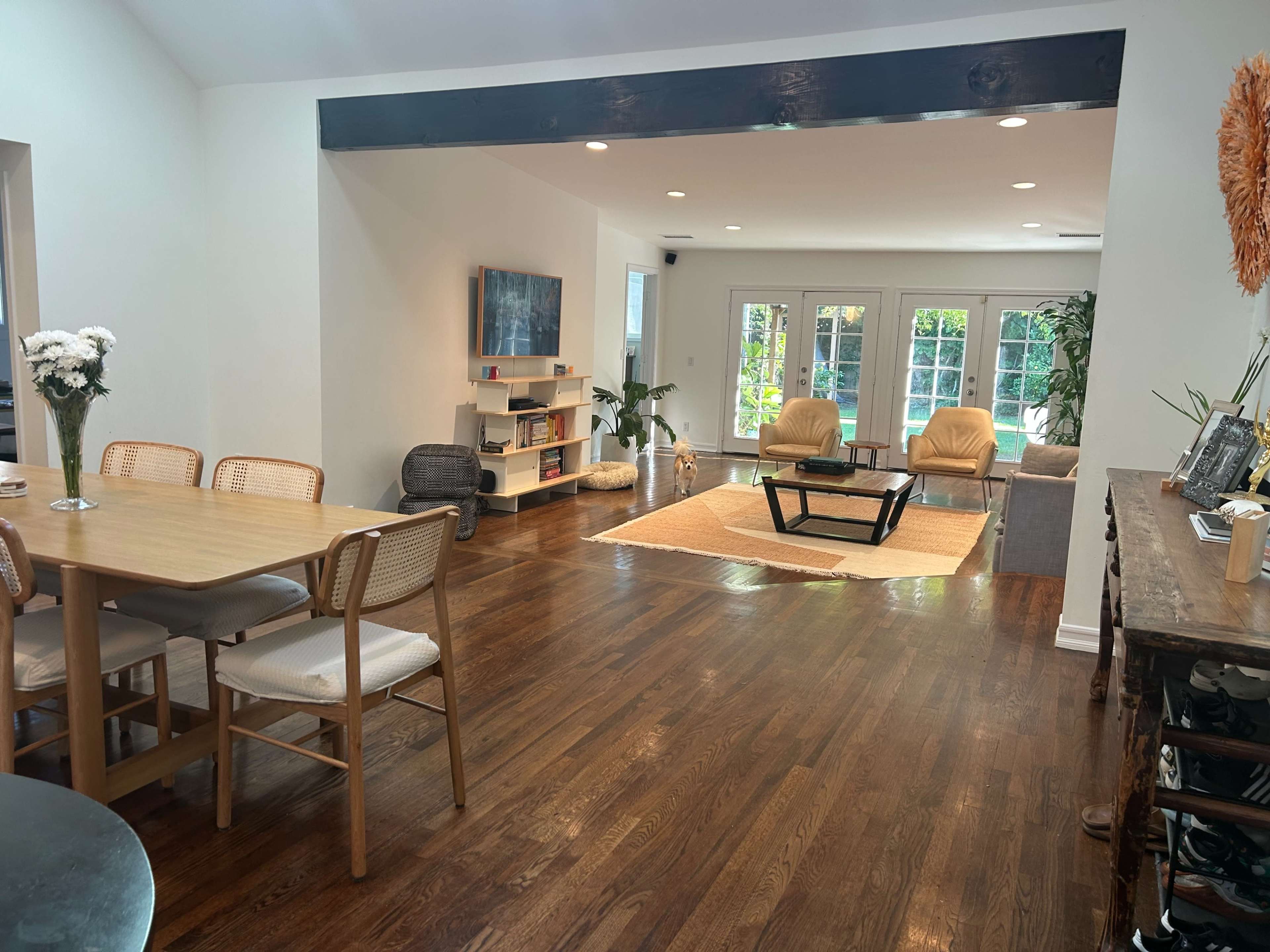 The image shows a modern interior space featuring a dining area with a wooden table and chairs, leading into a living room with two armchairs and large windows letting in natural light.