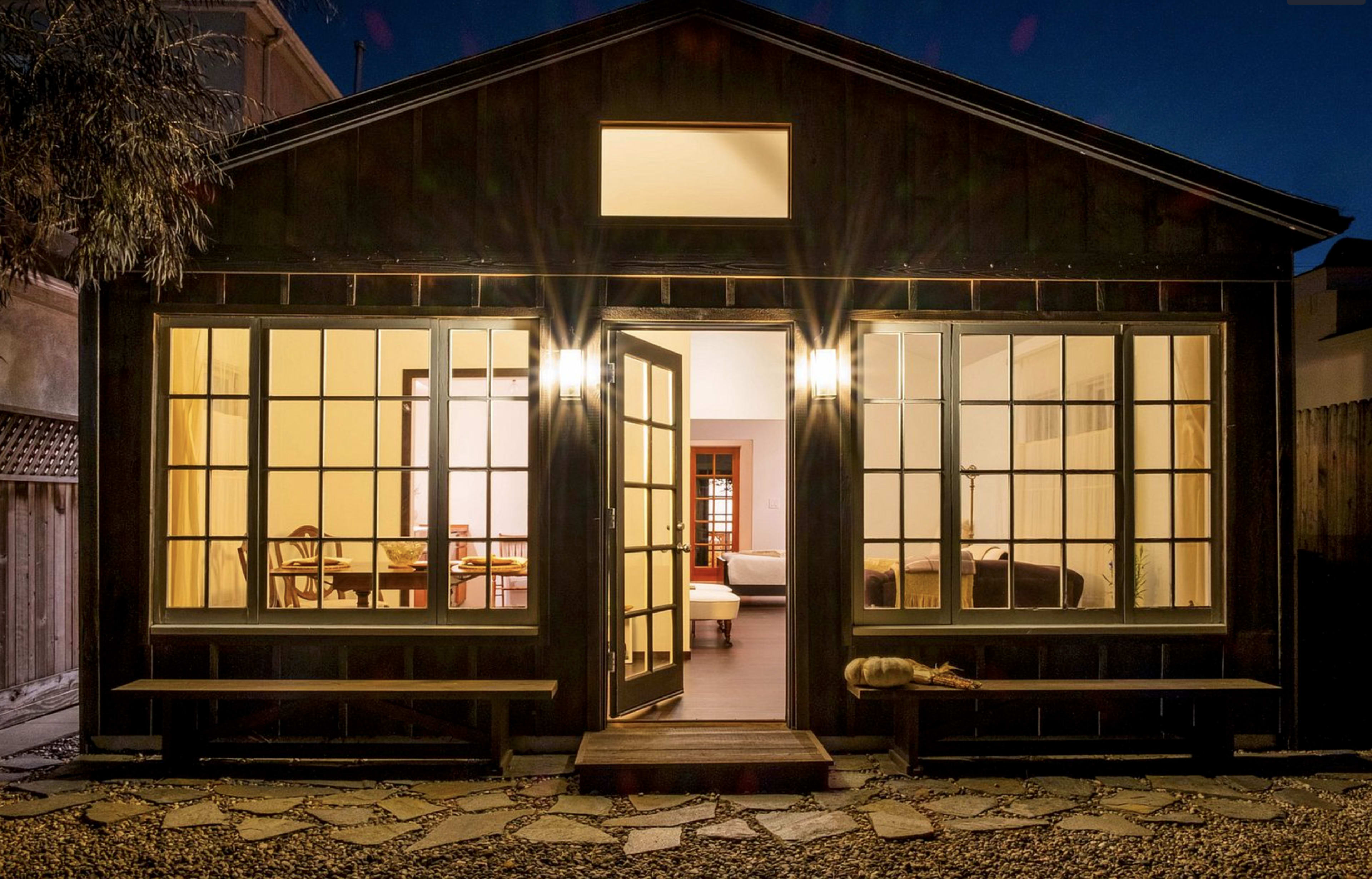 A dark wooden house with large front windows is lit from within, revealing a dining area and a bedroom, with a stone path leading to the entrance.