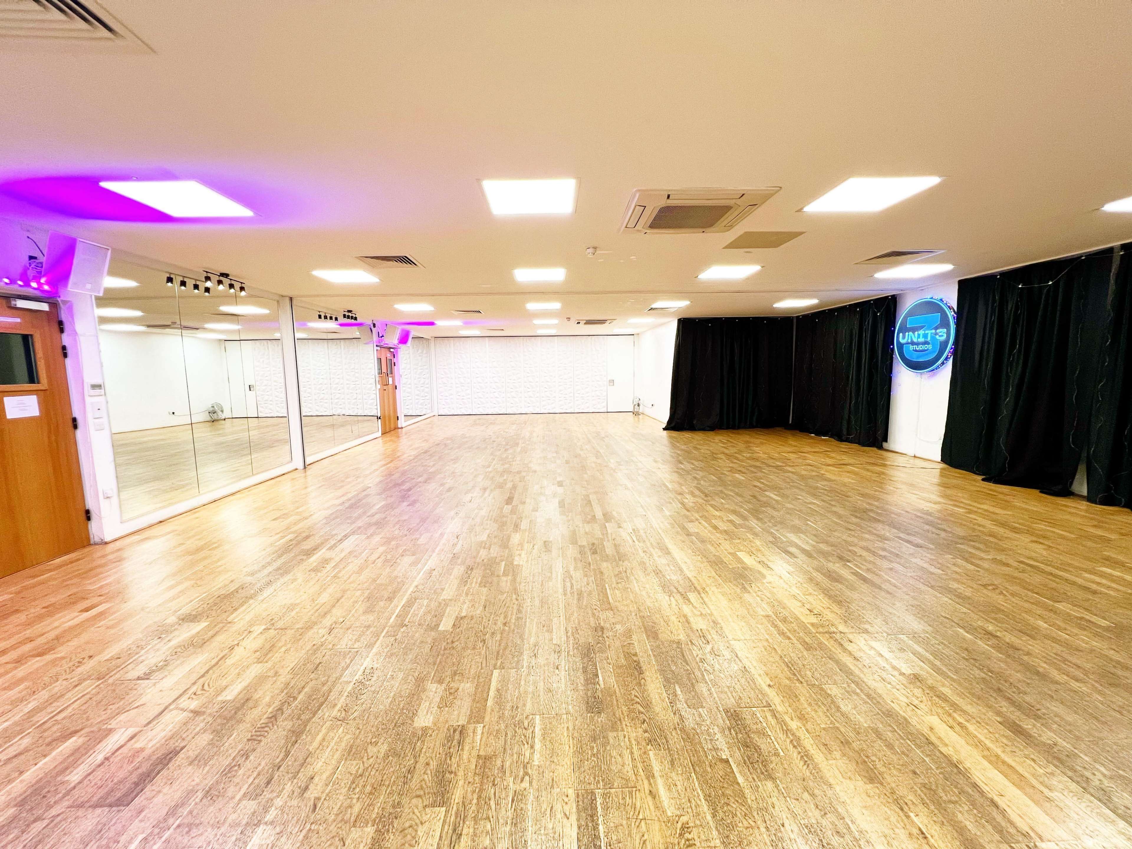 The image shows a spacious, well-lit dance studio with wooden flooring and mirrors along one wall.