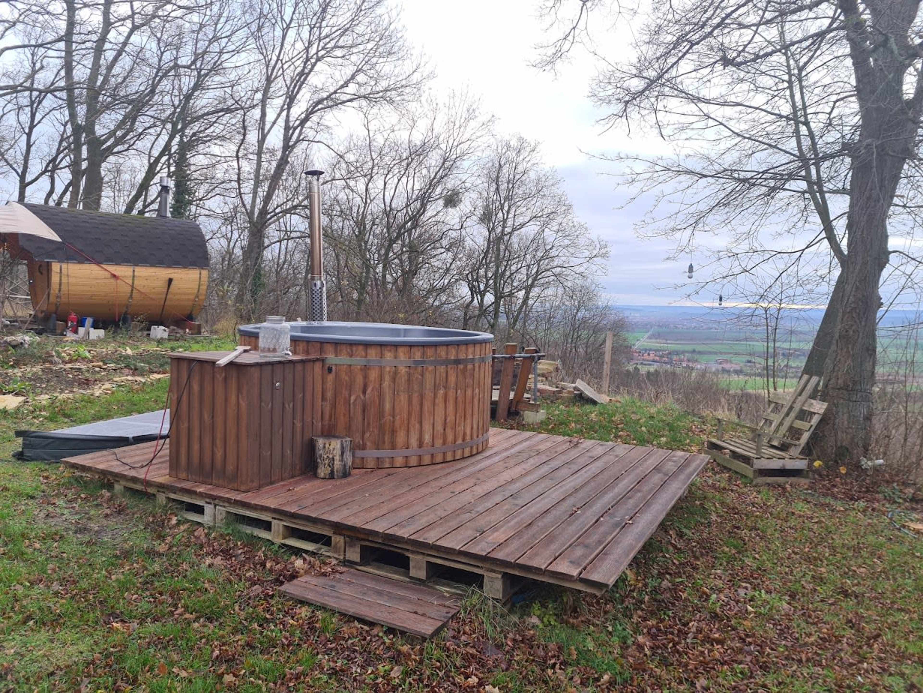 A wooden hot tub is situated on a deck surrounded by trees, overlooking a valley.
