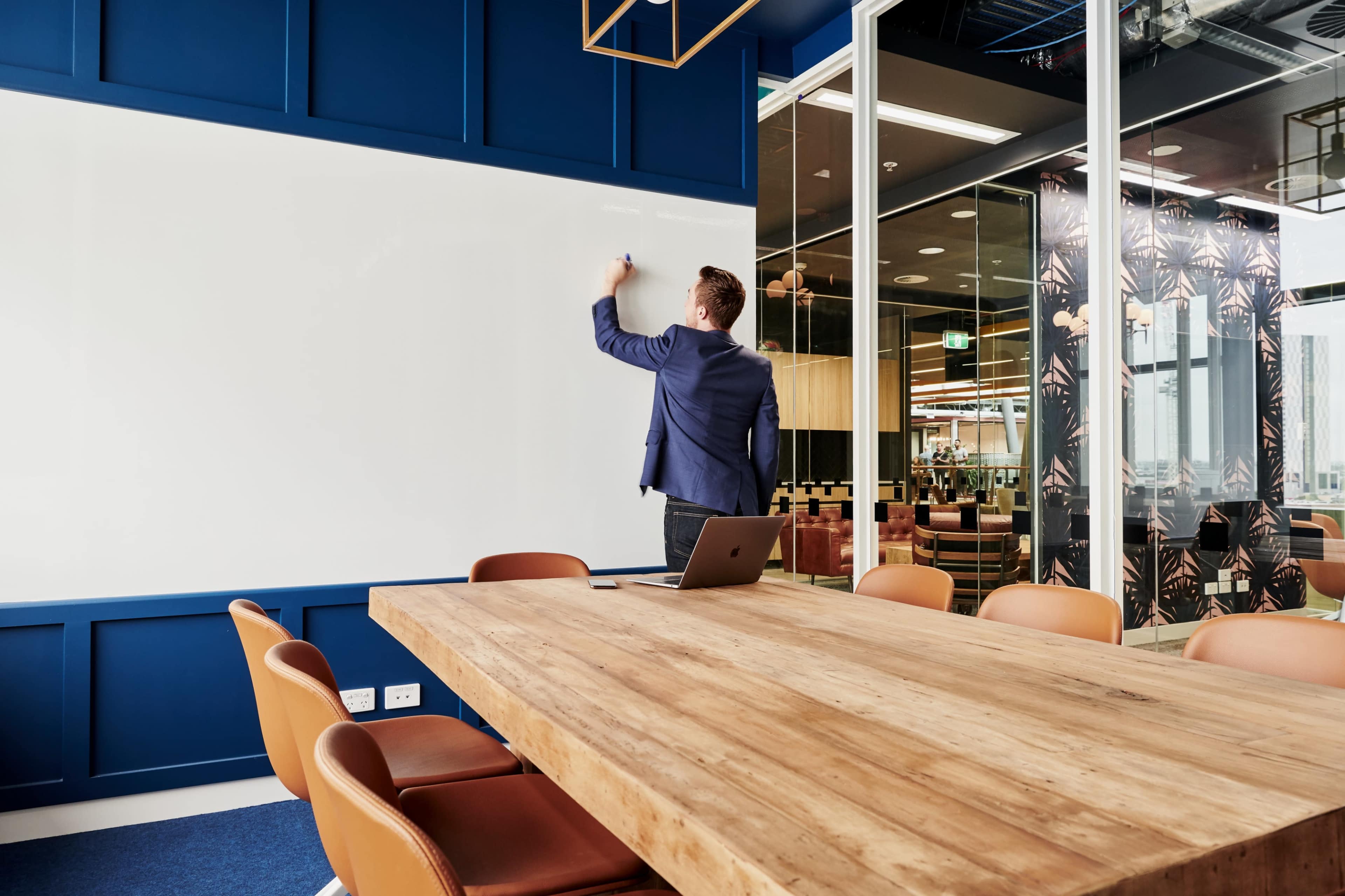 A man in a suit writes on a large whiteboard in a modern conference room with a wooden table and glass walls.