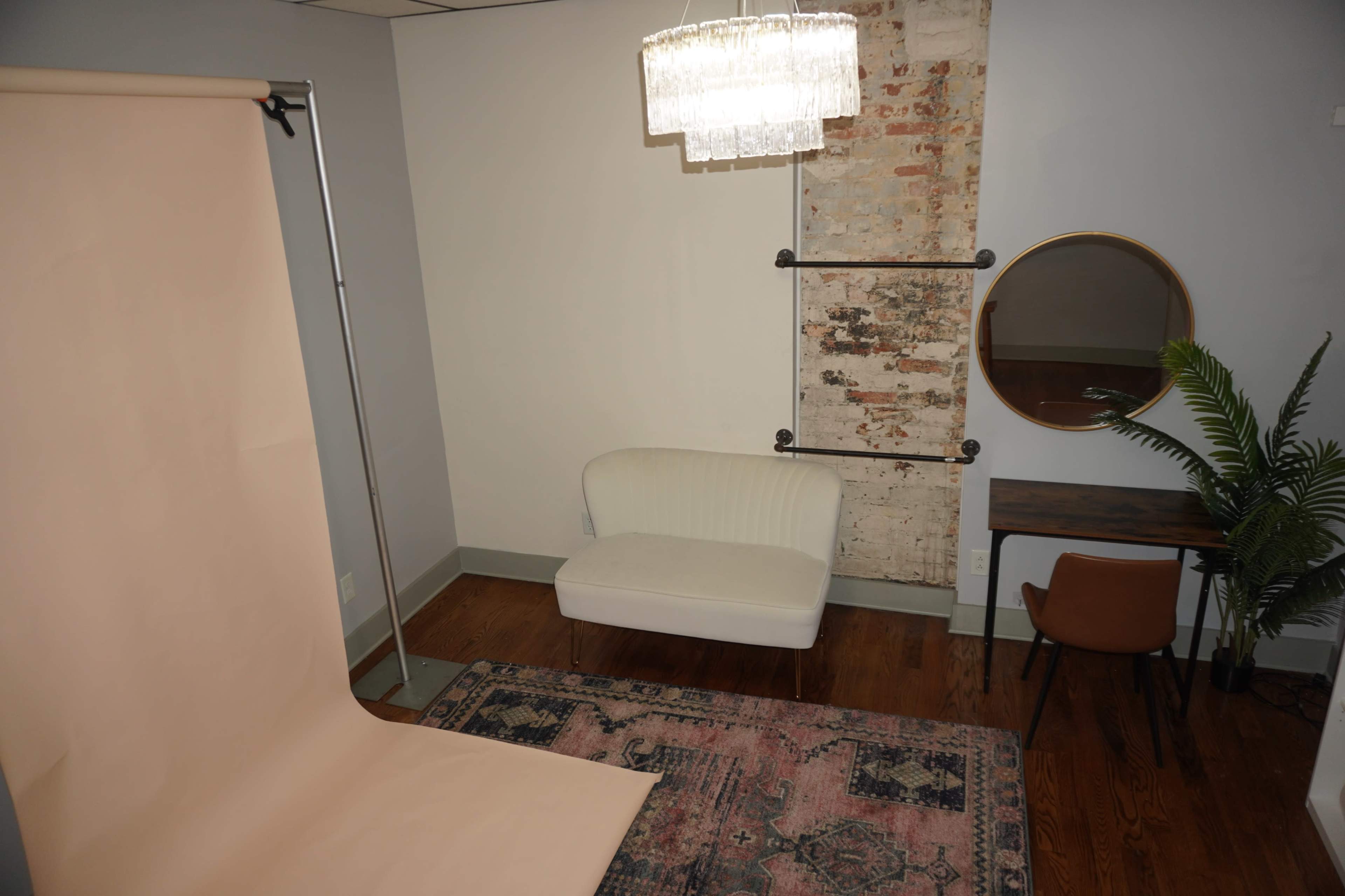 The image shows a small room with a cream-colored backdrop, a white sofa, a small wooden desk, a round mirror, and a decorative plant, all against a brick wall.