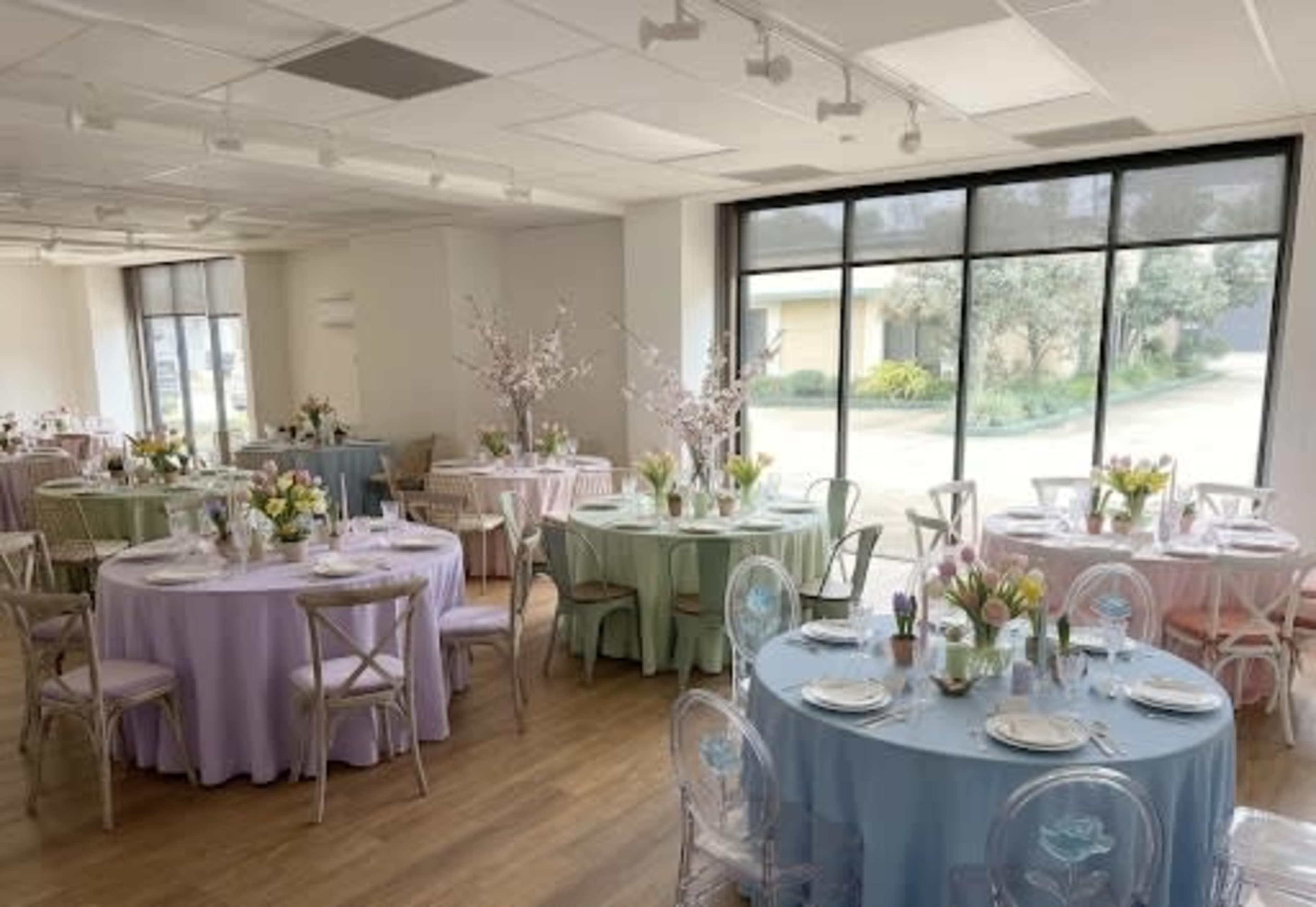 The image shows a bright room with several round tables, each covered in pastel tablecloths, arranged for a dining event with floral centerpieces.