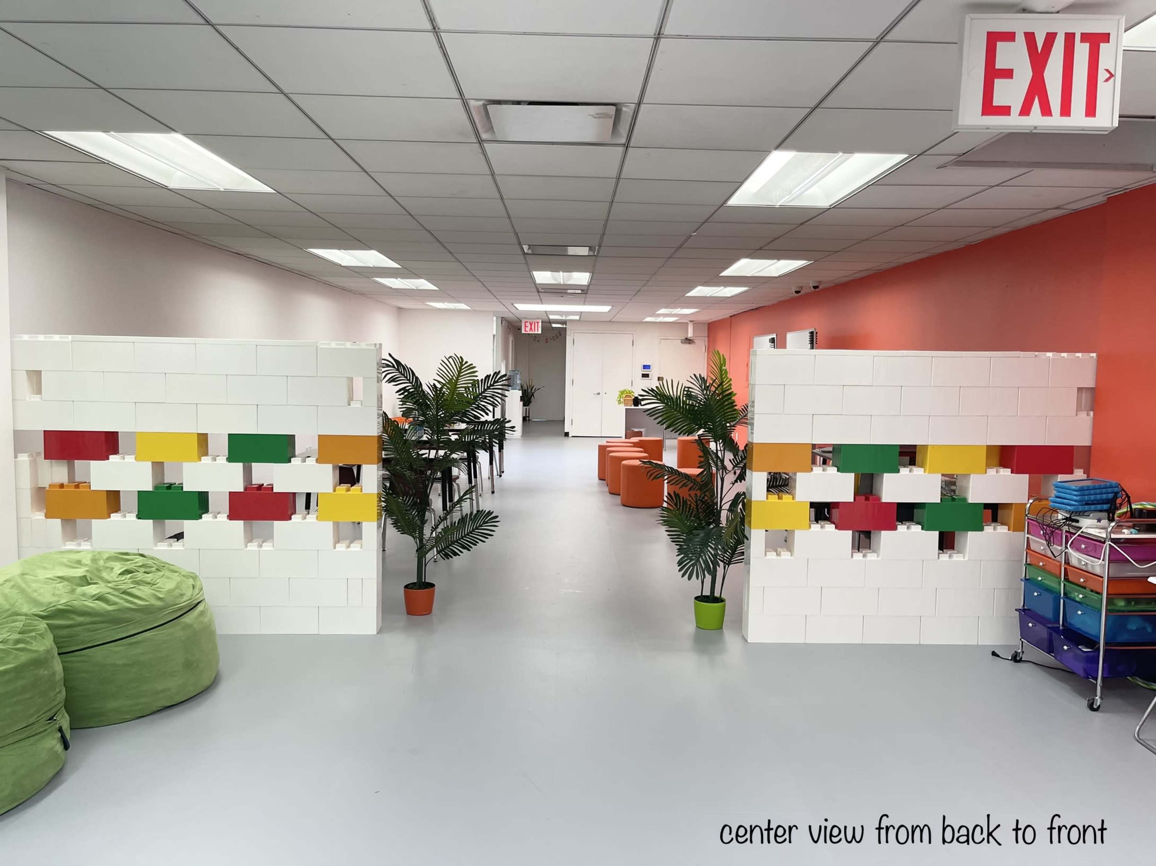 A brightly lit interior hallway with two colorful partition walls made of blocks, flanked by potted plants and bean bags, leading to an exit sign at the far end.