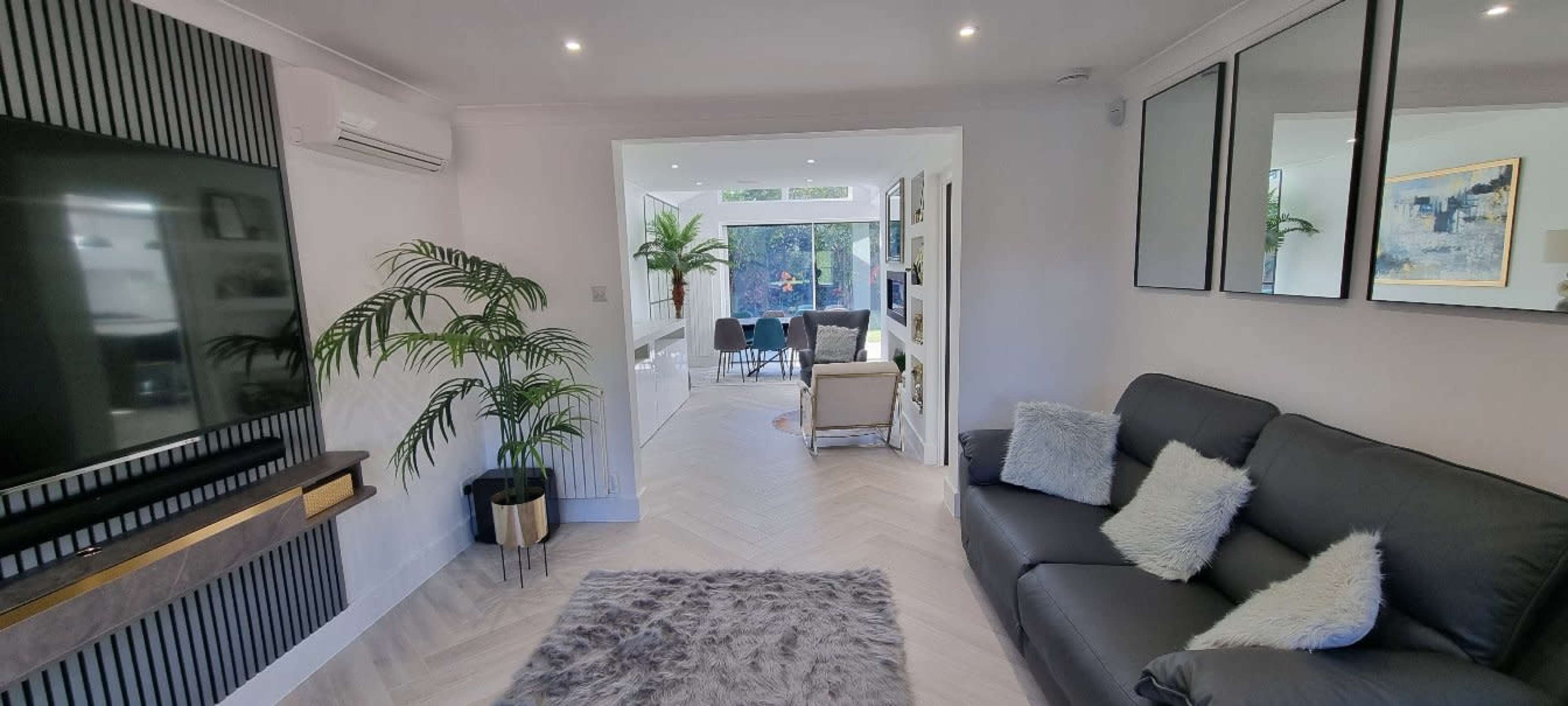 A modern living room features a leather sofa, a television on a wall-mounted unit, and a view of an adjoining space with plants and seating.