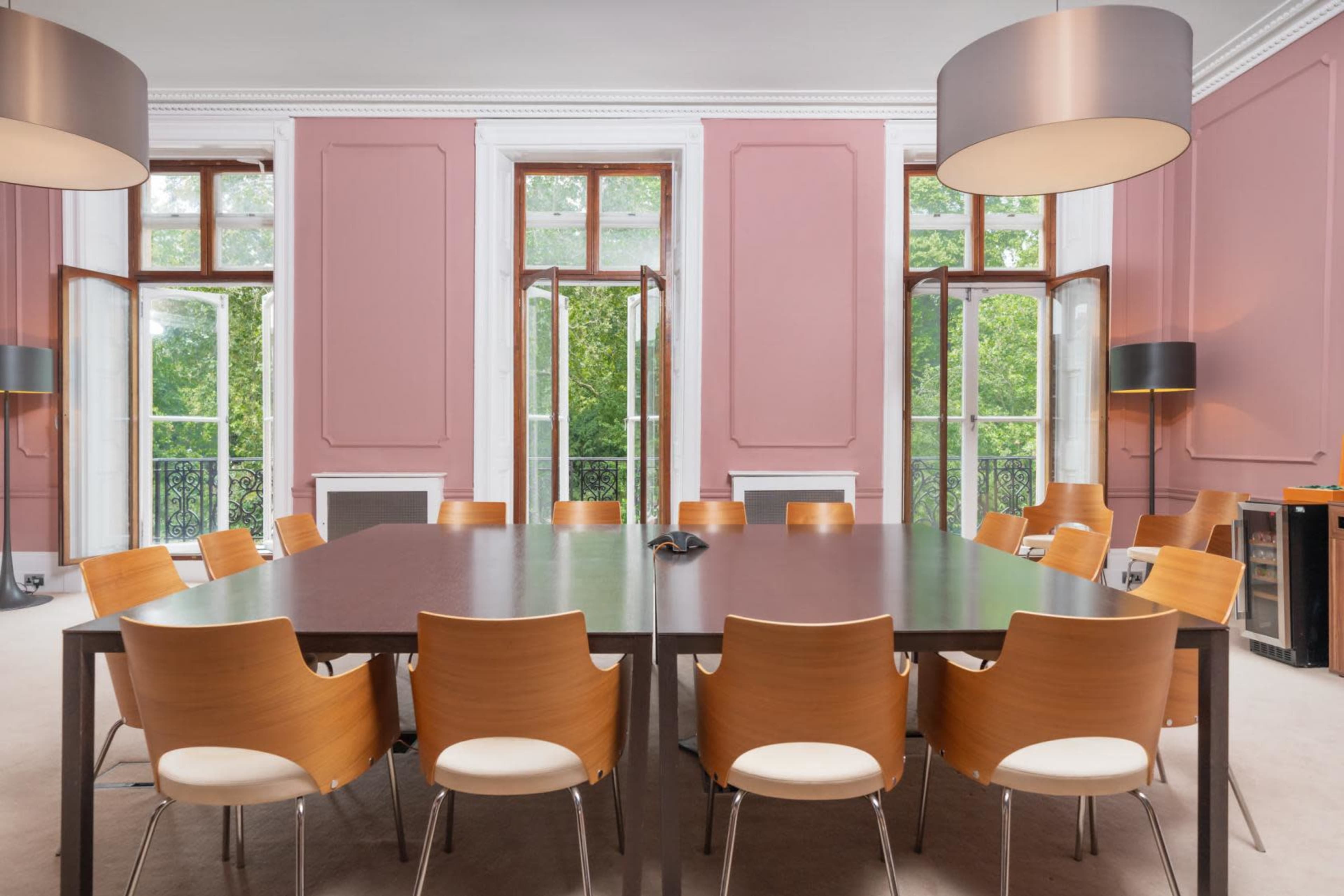 A large conference room features a central table surrounded by wooden chairs, with tall windows allowing natural light and a view of greenery outside.