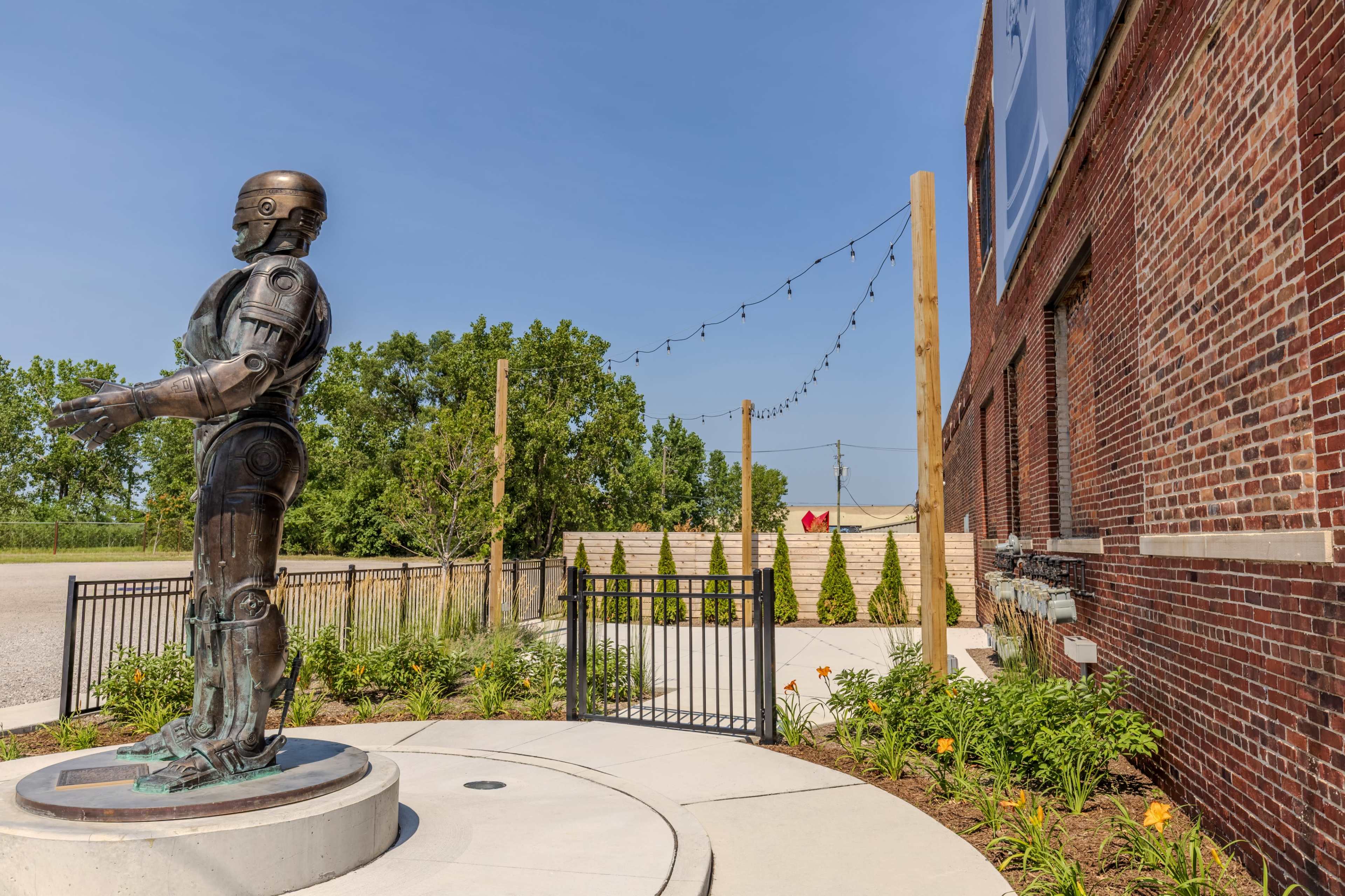 A metallic robot sculpture stands on a circular concrete base near a brick wall, surrounded by landscaped greenery and string lights overhead.