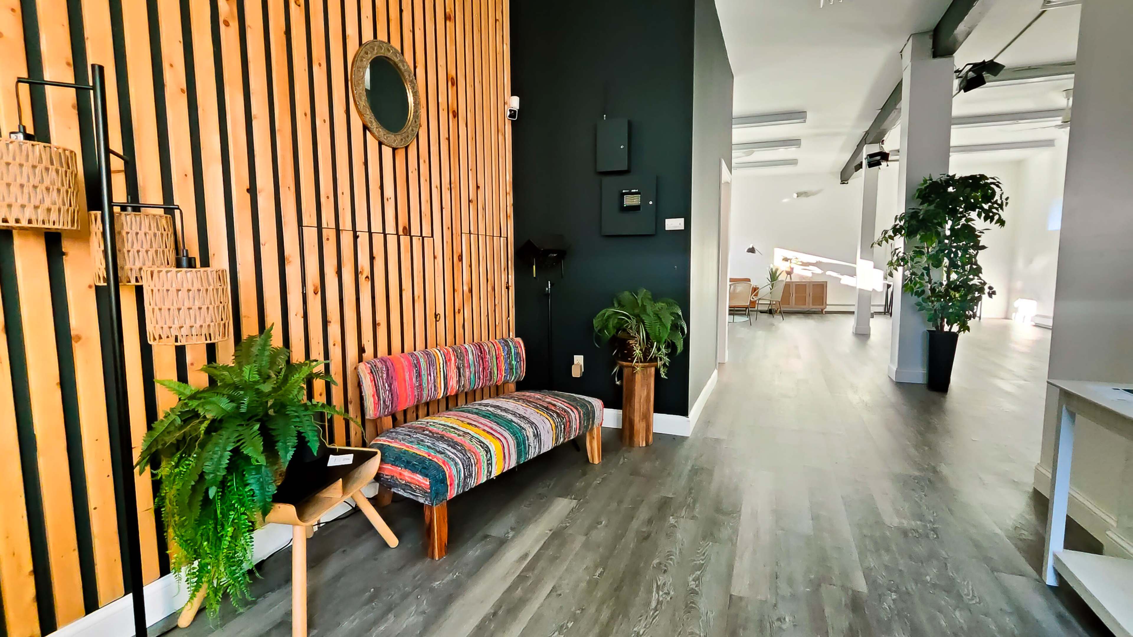 A brightly lit interior features a wooden wall, a colorful striped sofa, and green plants, leading to an open area with contemporary decor.