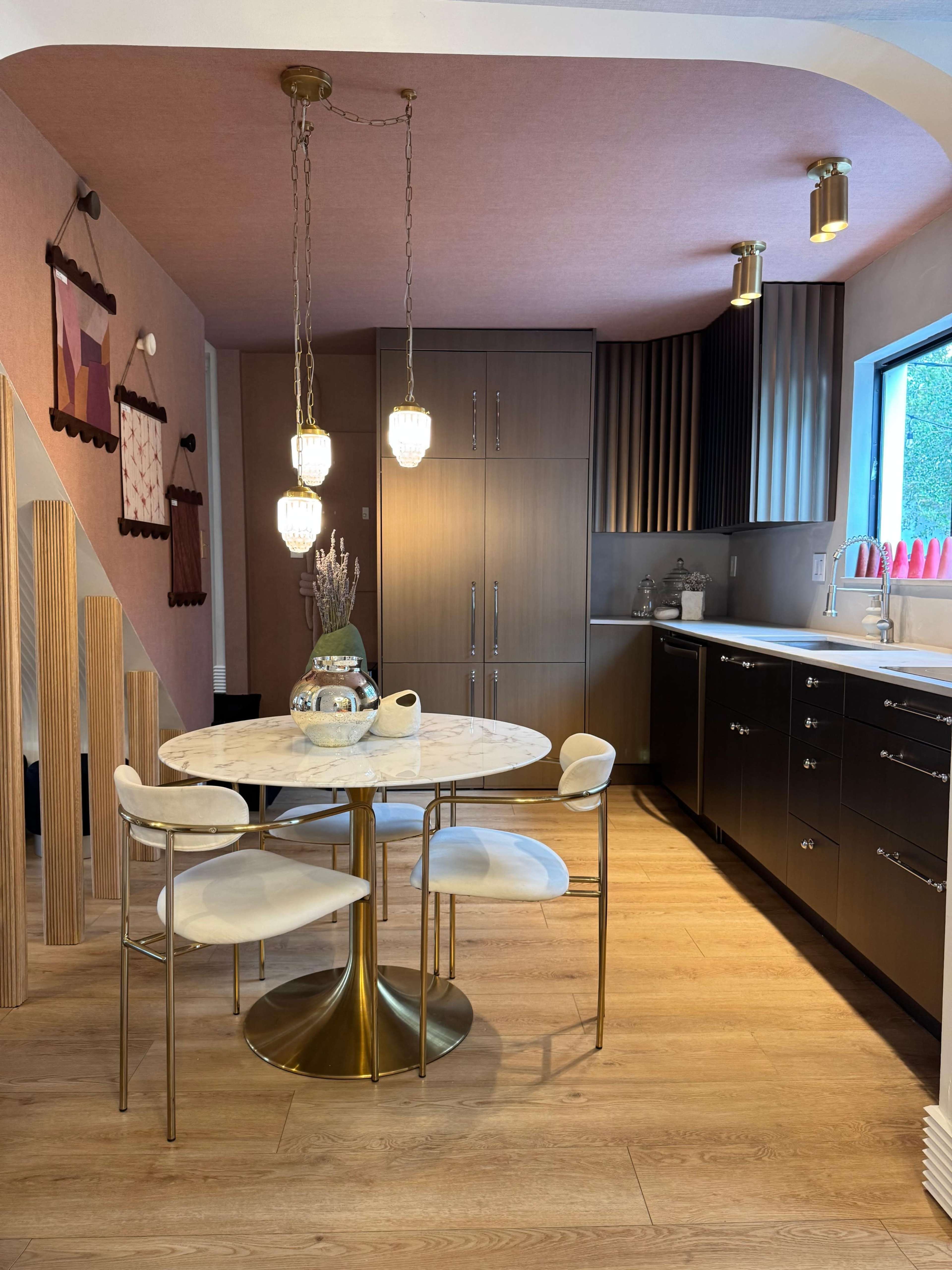 The image shows a modern kitchen with a round marble dining table surrounded by four white chairs, pendant lights hanging from the ceiling, and cabinetry in varying shades of brown and black.