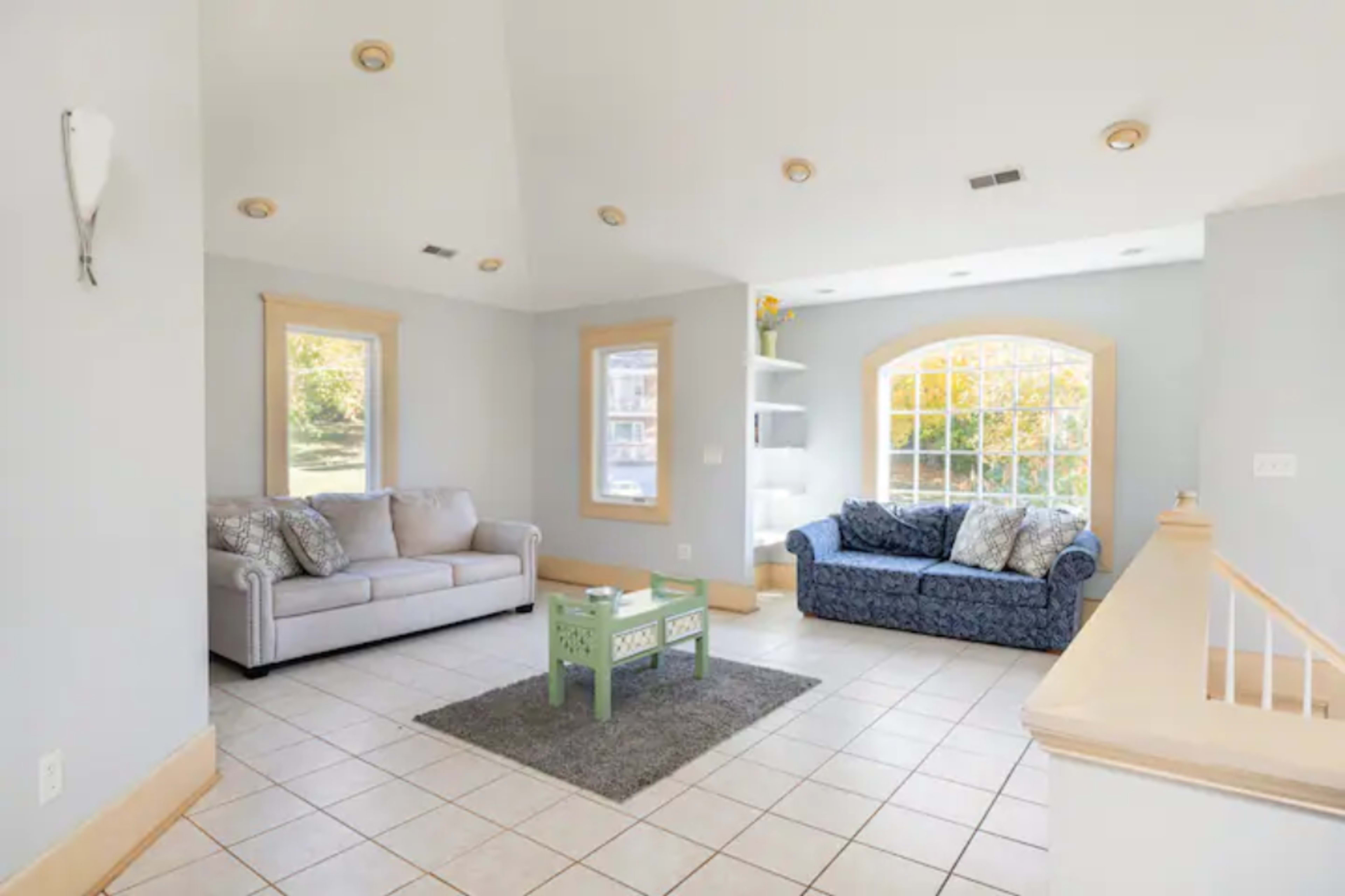 A bright living room features two couches, a green coffee table, and large windows allowing natural light.