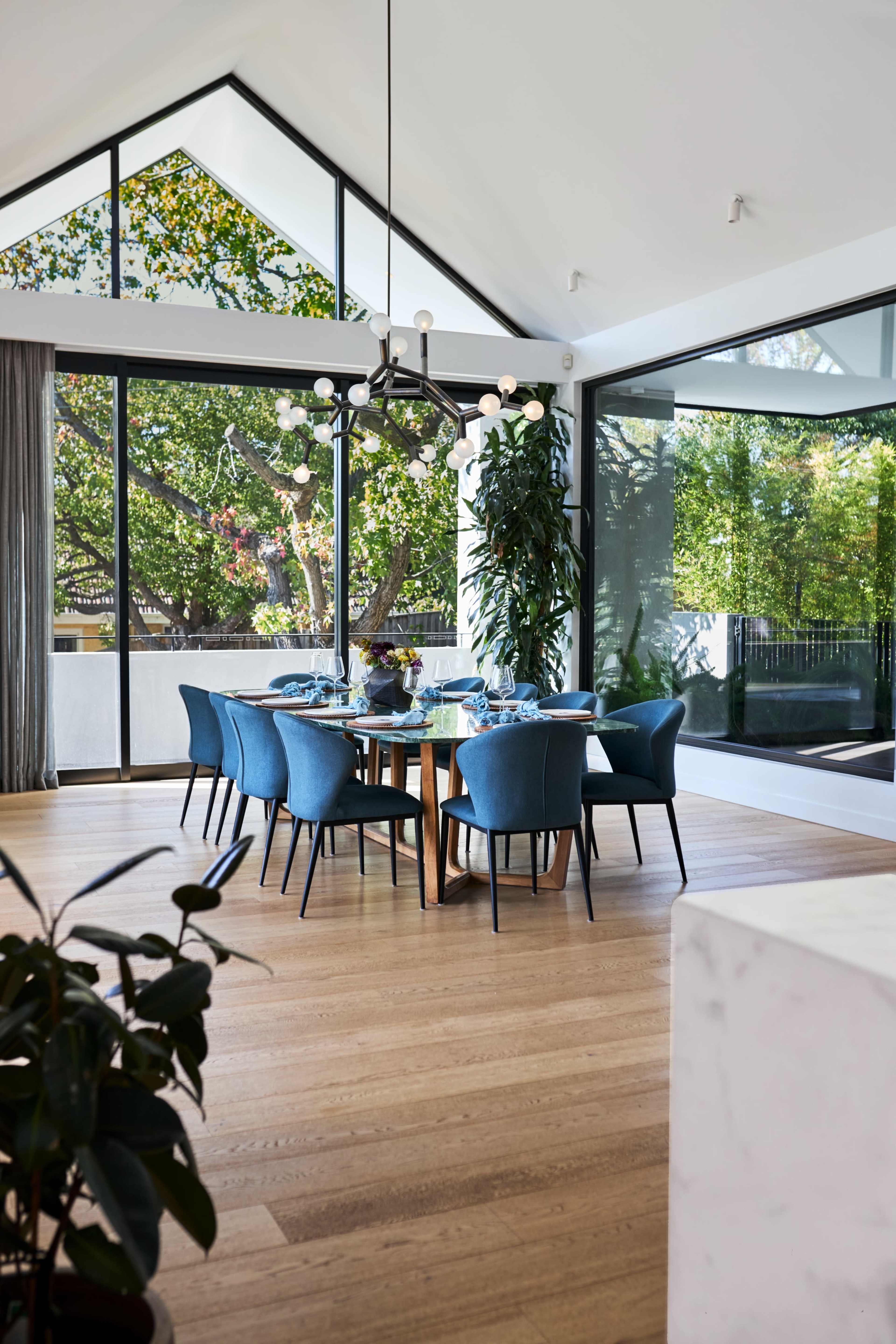 A modern dining room features a large wooden table surrounded by blue chairs, with tall windows showcasing greenery outside.