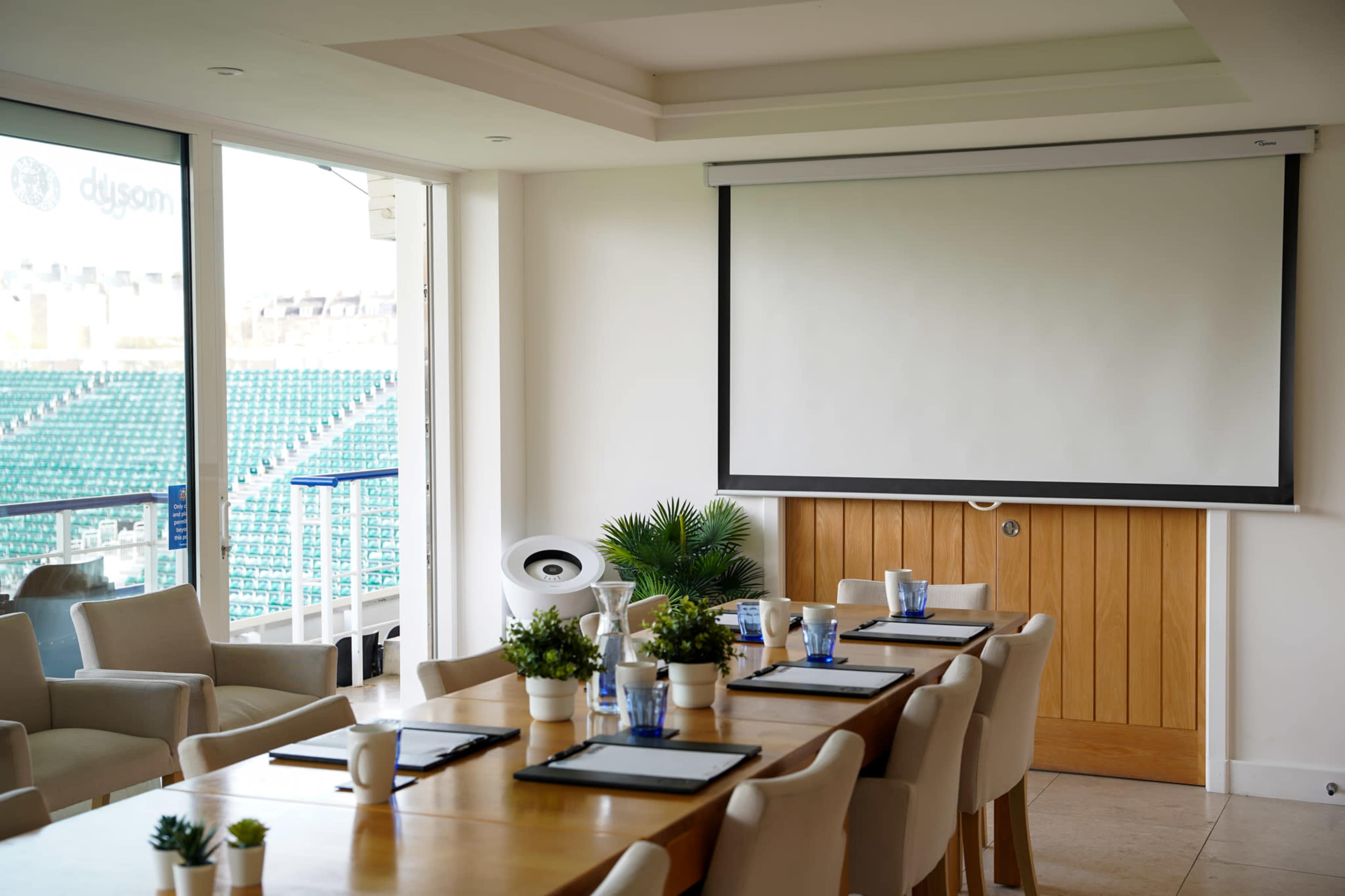 A modern meeting room features a large wooden table set with place settings, a projection screen on the wall, and views of an empty stadium through large windows.