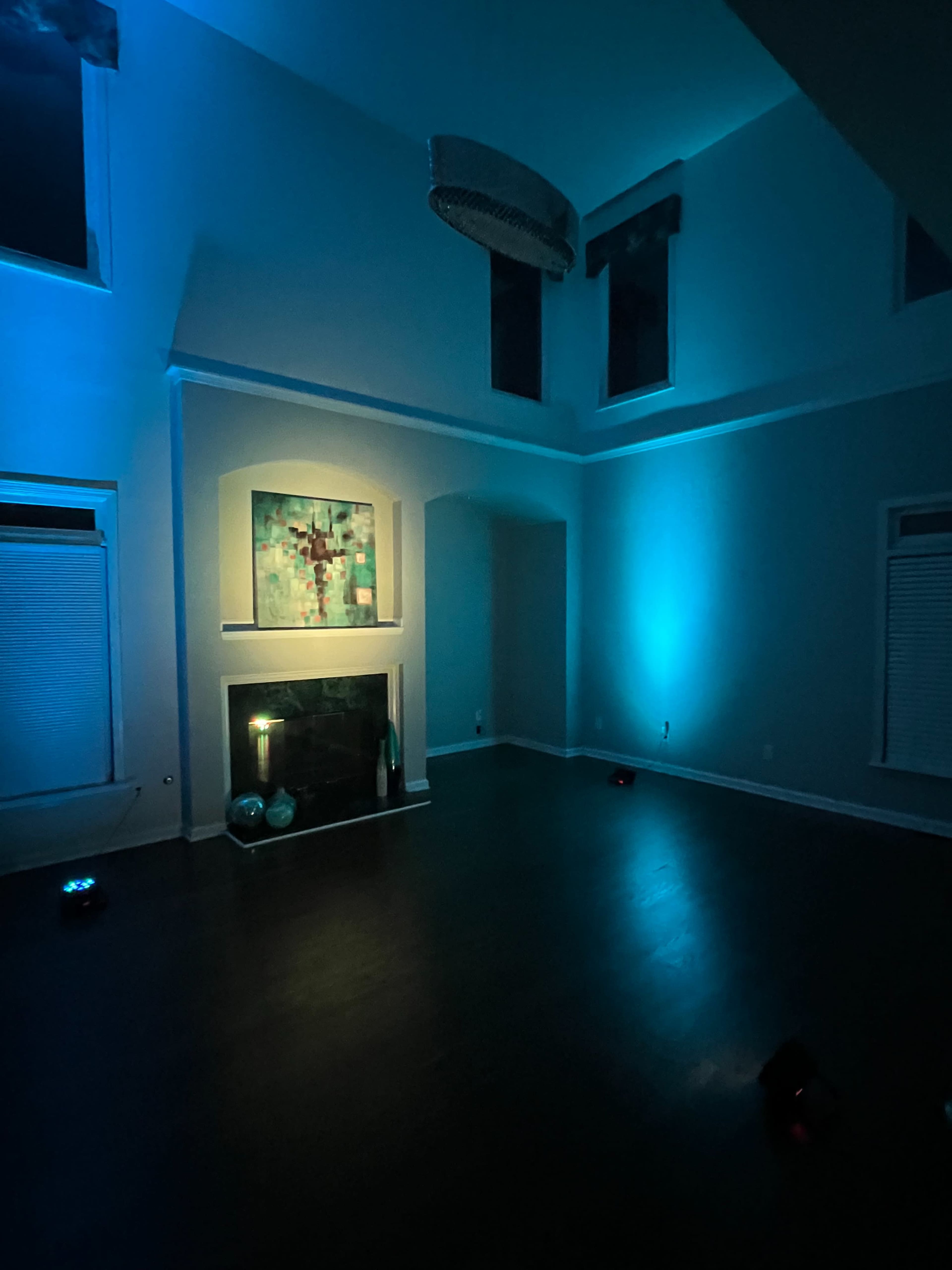 A dimly lit room with a high ceiling features blue ambient lighting, an artwork above a fireplace, and a dark wooden floor.