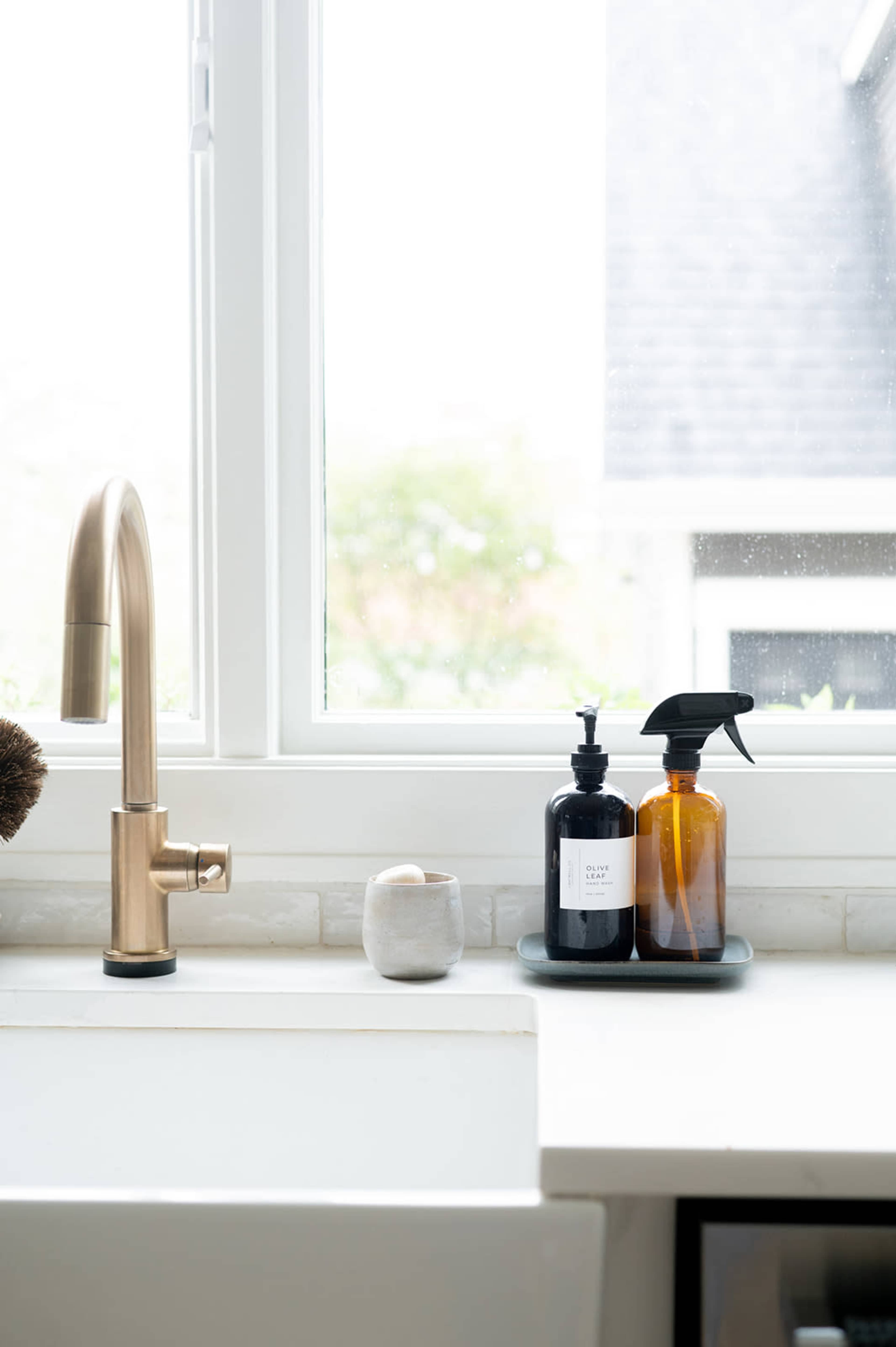 A modern kitchen sink area features a bronze faucet, two dark bottles on a tray, and a white ceramic vessel, with natural light streaming through a nearby window.