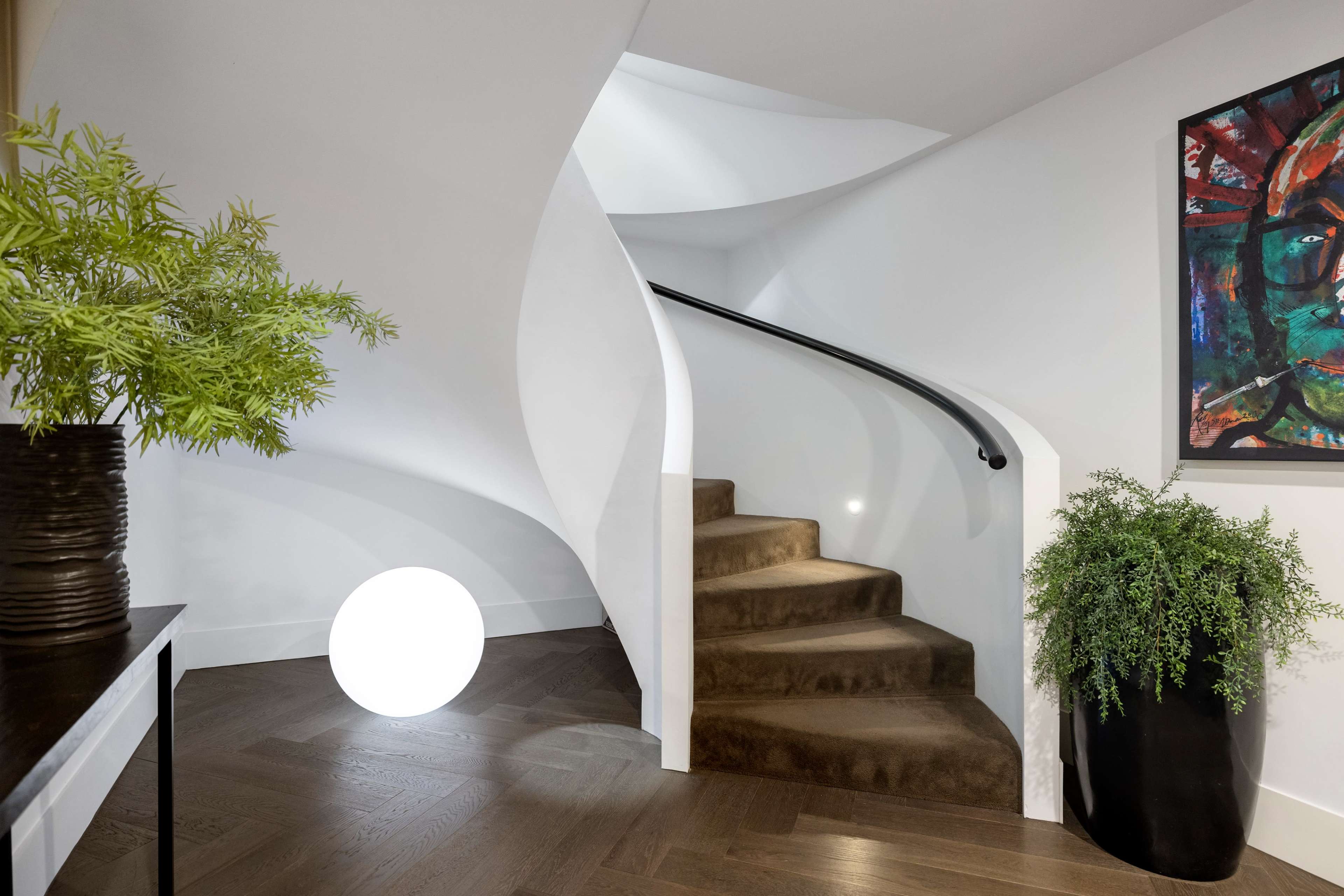 A curved staircase with a dark railing leads up from a light-colored entryway, accompanied by a large round light fixture and potted plants.