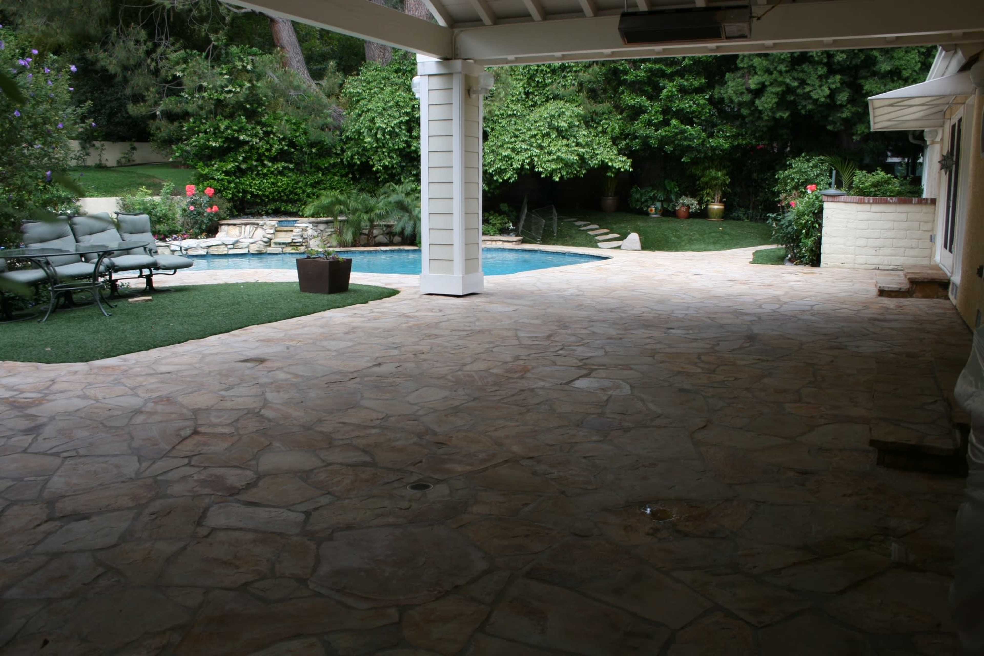 The image shows a spacious patio area with a stone floor, leading to a swimming pool surrounded by greenery.