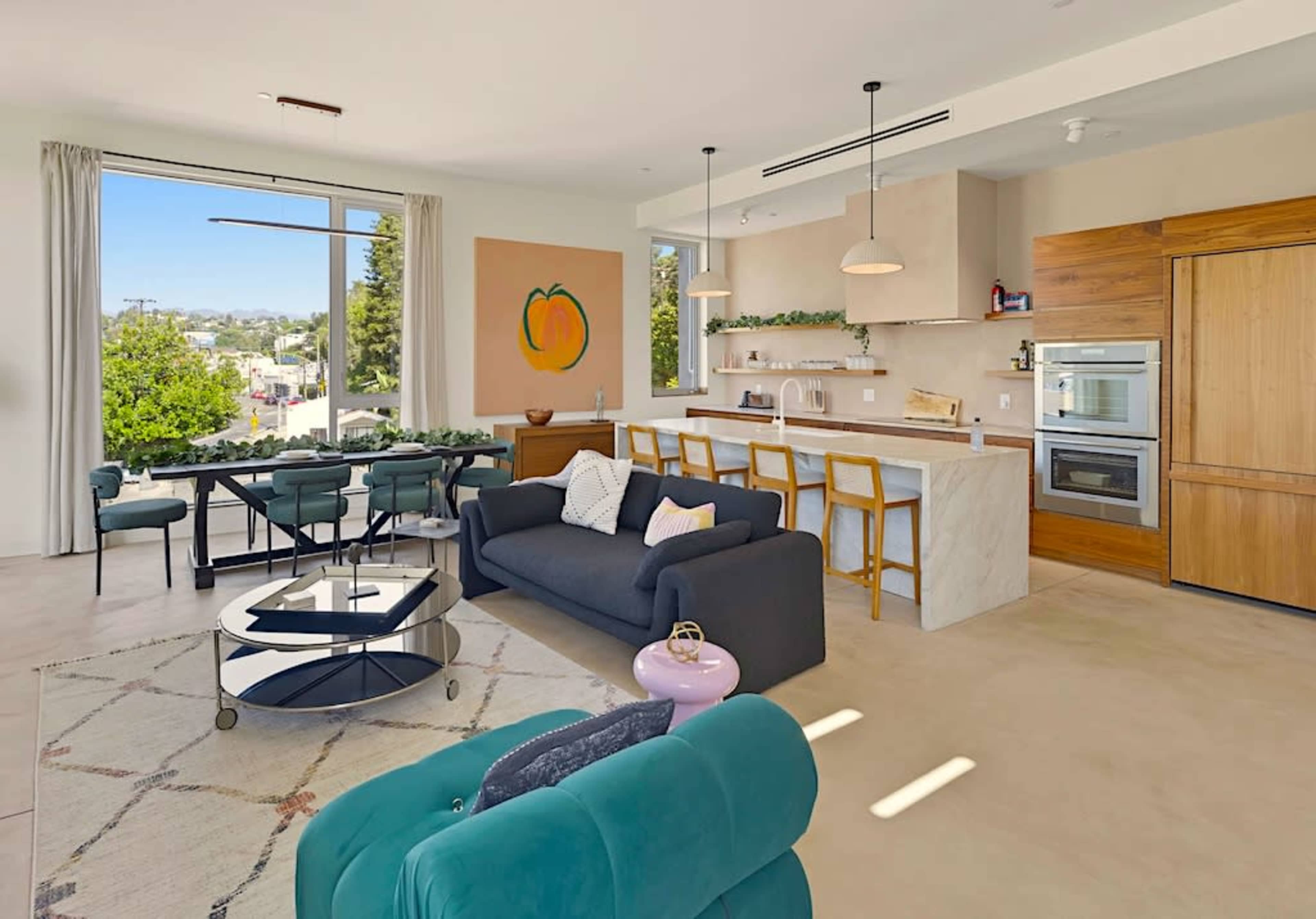 The image shows a modern living space with a kitchen, dining area, and a comfortable seating arrangement, featuring large windows that provide natural light and a view of the outdoors.