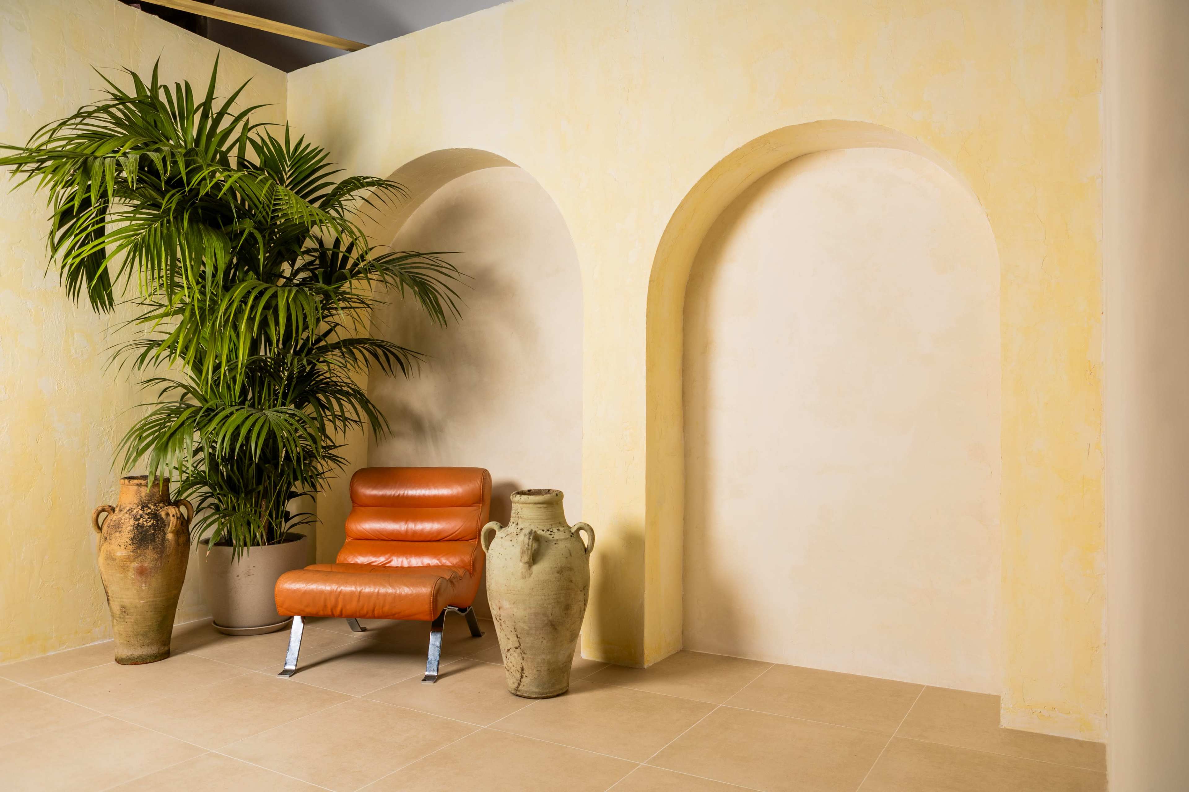 The image features a minimalist interior with a palm plant, a modern orange chair, and two decorative clay pots against a softly colored wall.