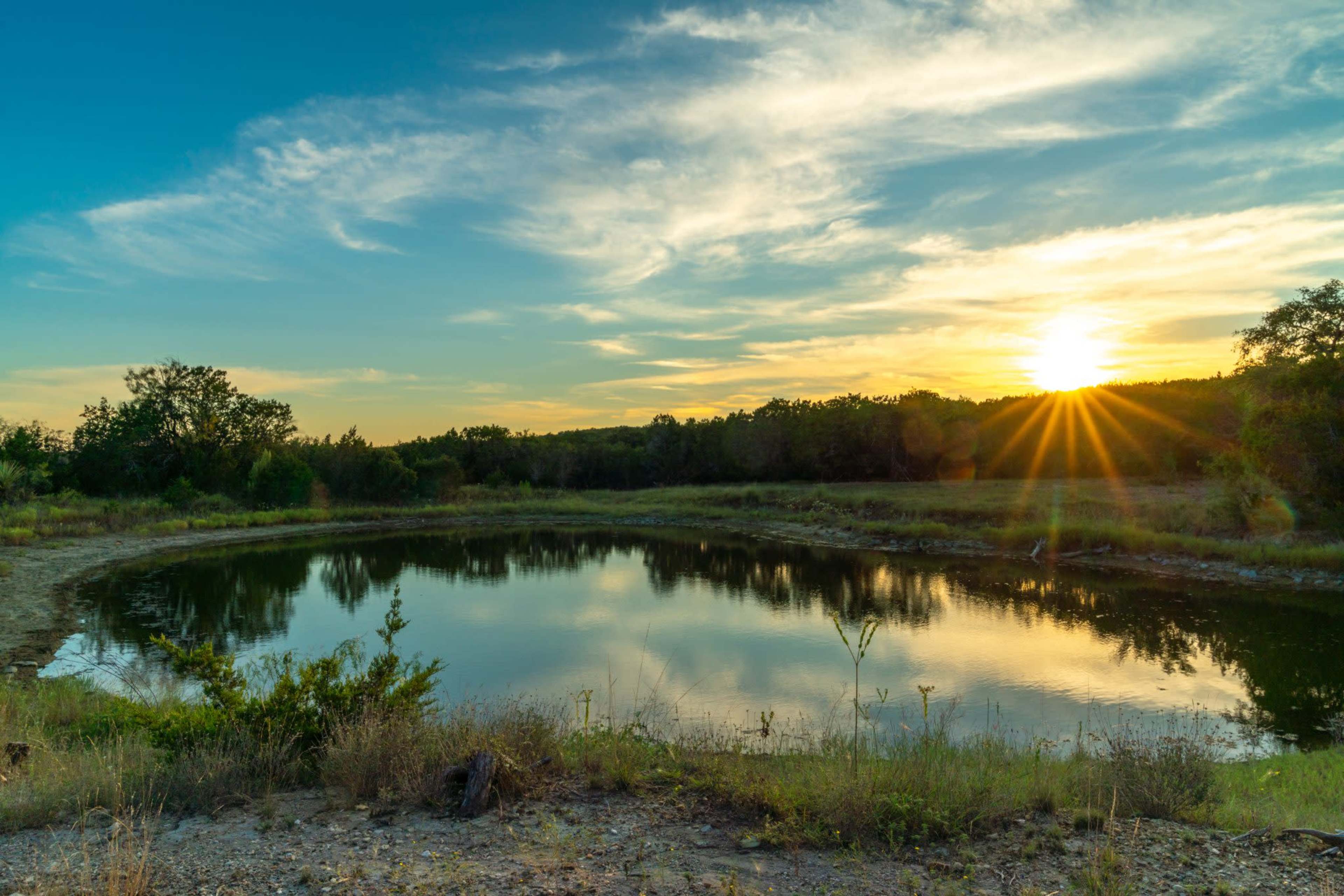 The sun sets over a calm pond, reflecting the sky and surrounding trees.