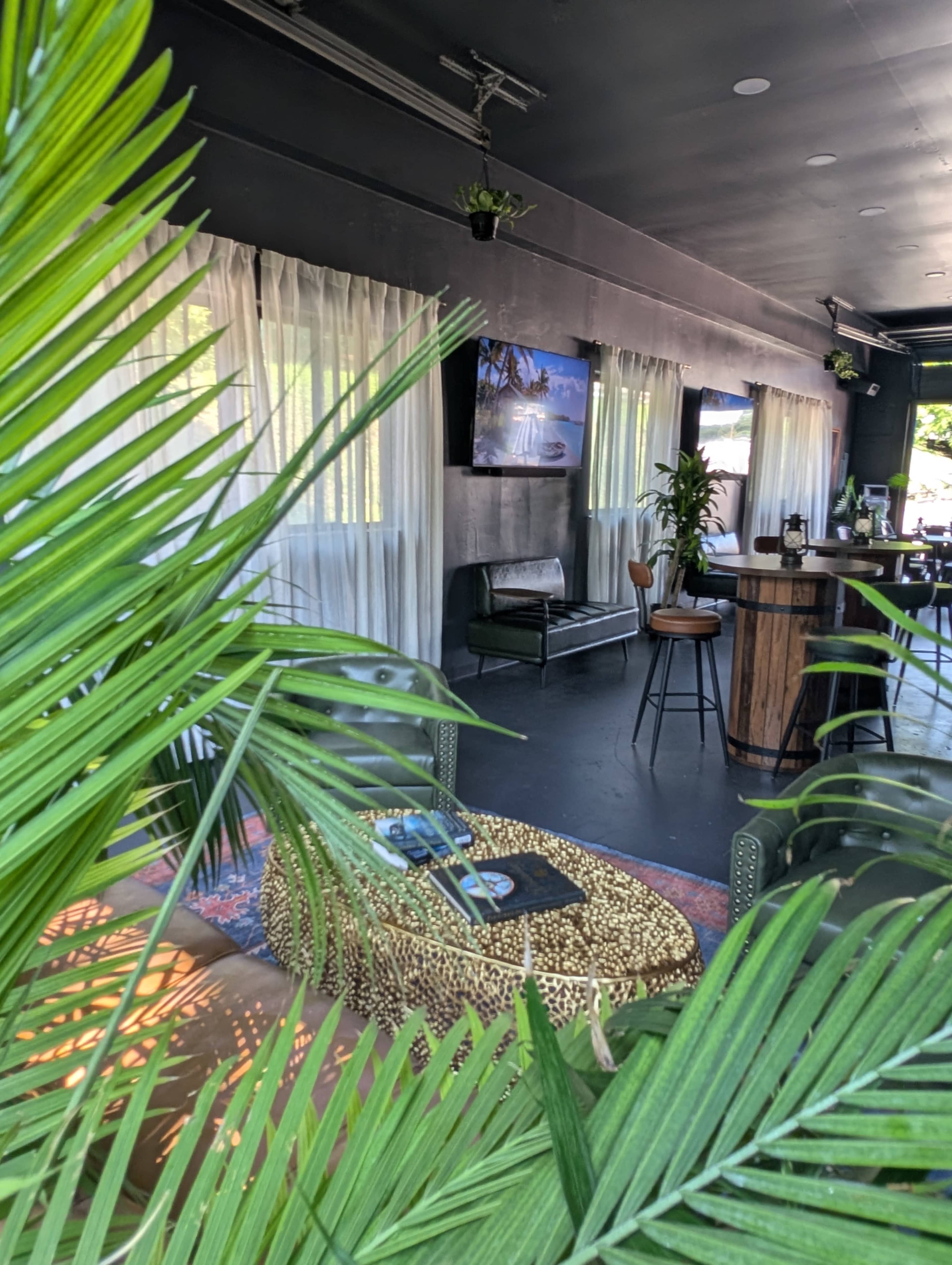 The image shows a stylish indoor bar area with plants, a seating area, and two screens displaying images.
