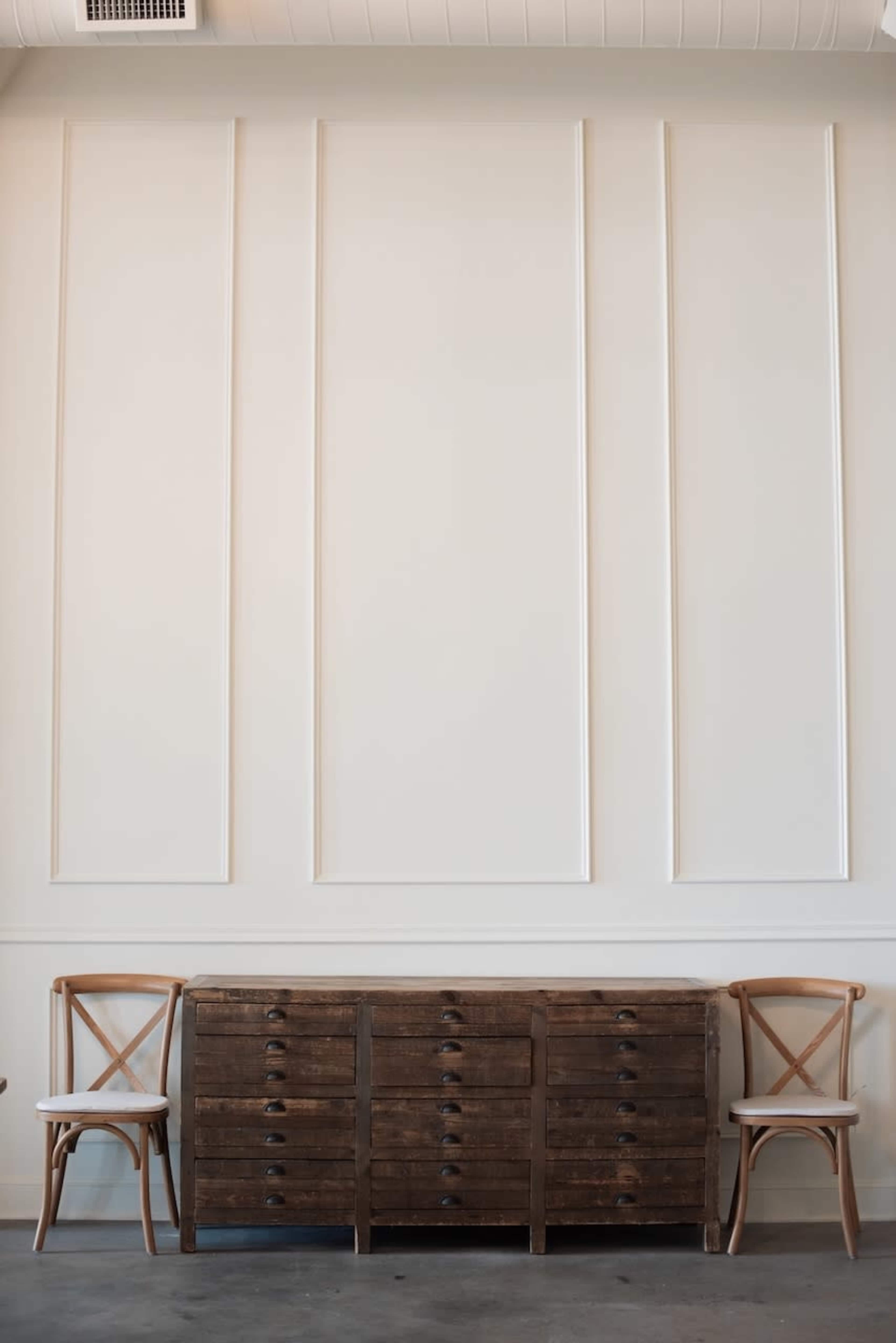 A wooden sideboard is positioned against a white paneled wall, flanked by two amber-colored chairs.