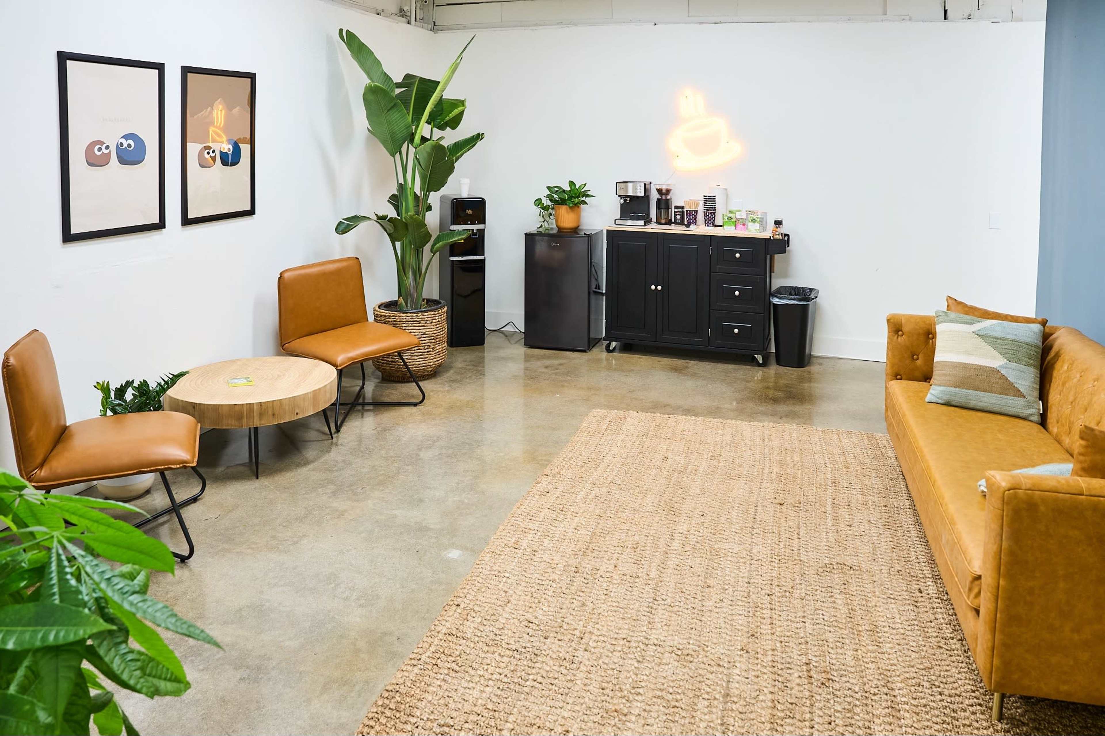 A modern lounge area featuring a brown sofa, two leather chairs, a round coffee table, a small kitchenette, and a decorative plant against a white wall.