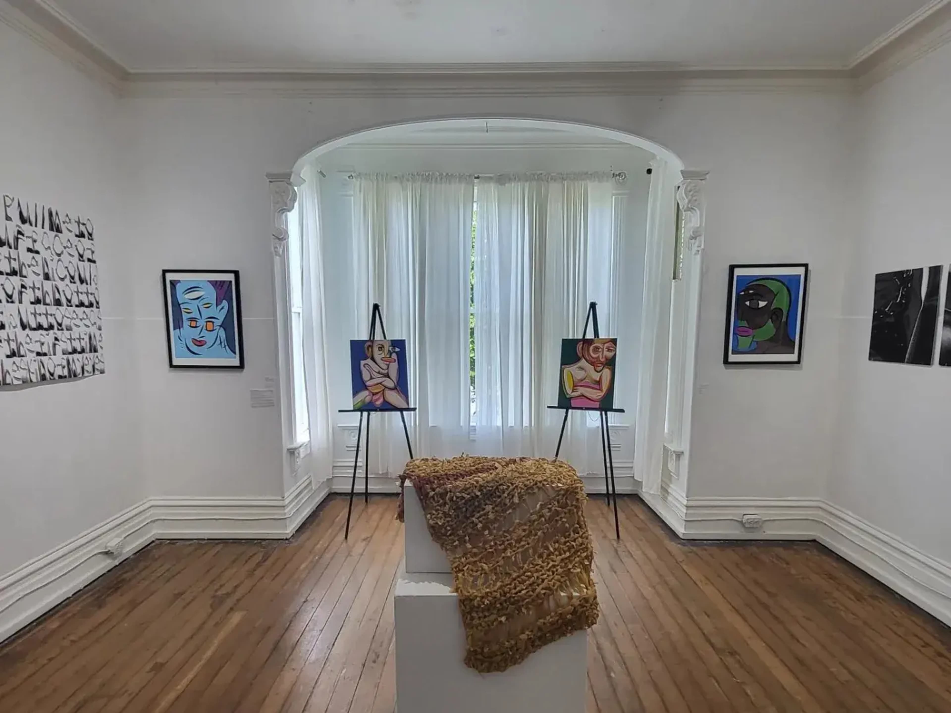 The image shows an art gallery with three paintings displayed on easels against a white wall, complemented by a textured piece on a pedestal.
