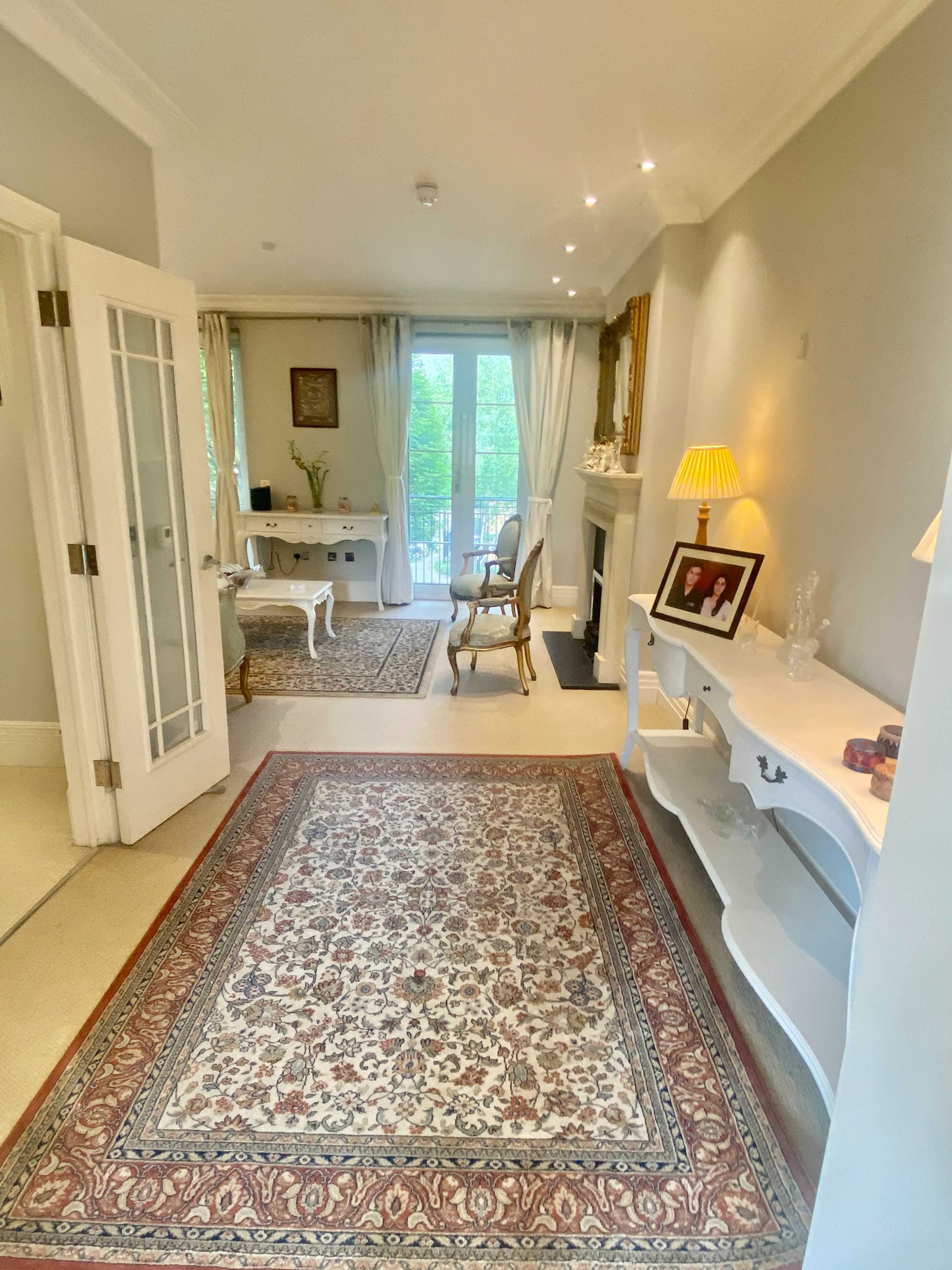 A well-decorated hallway features a patterned rug, a console table, and access to a bright room with a fireplace and large windows.
