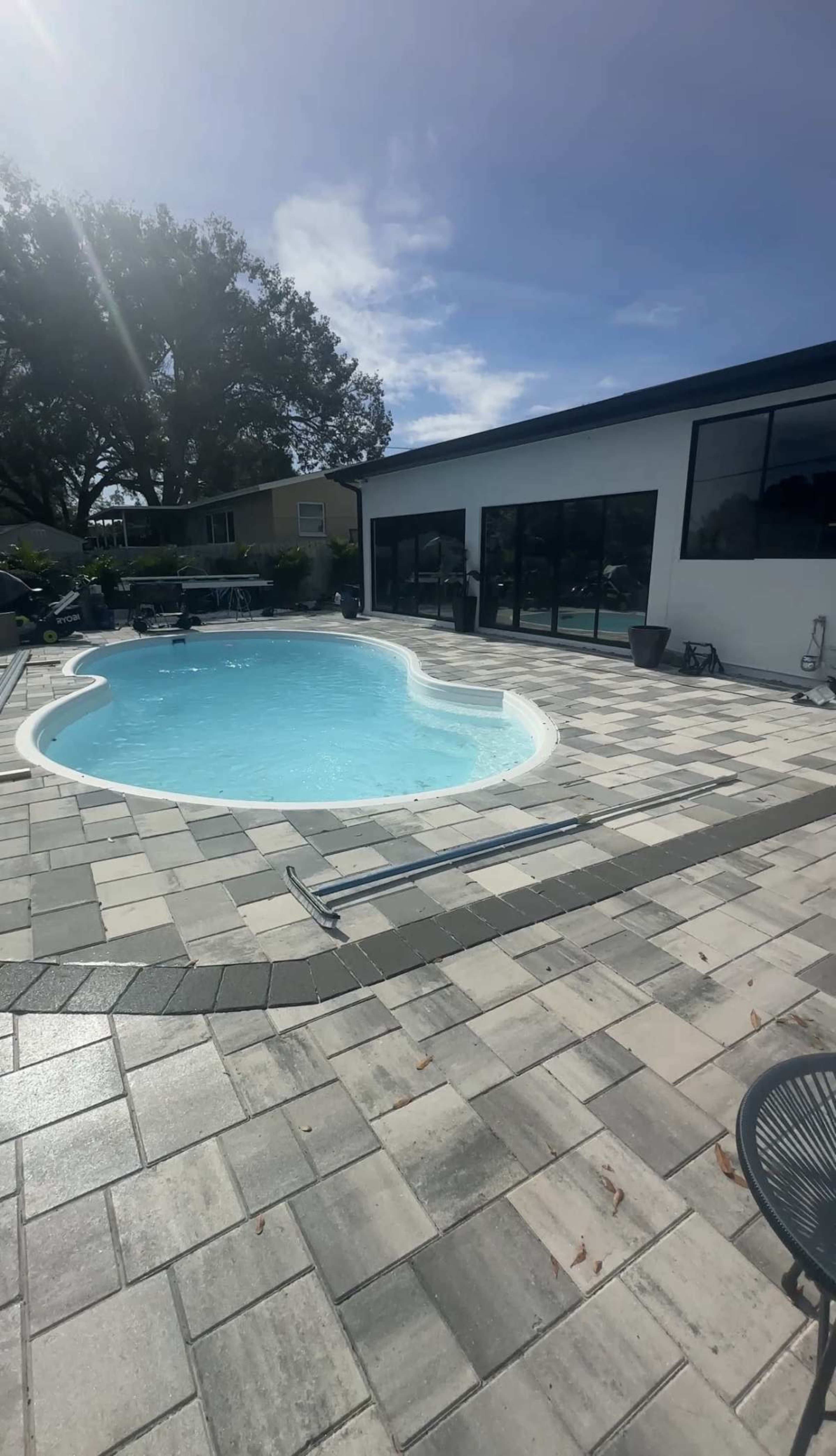 The image shows a swimming pool in a backyard surrounded by a paved area and a building with large windows.