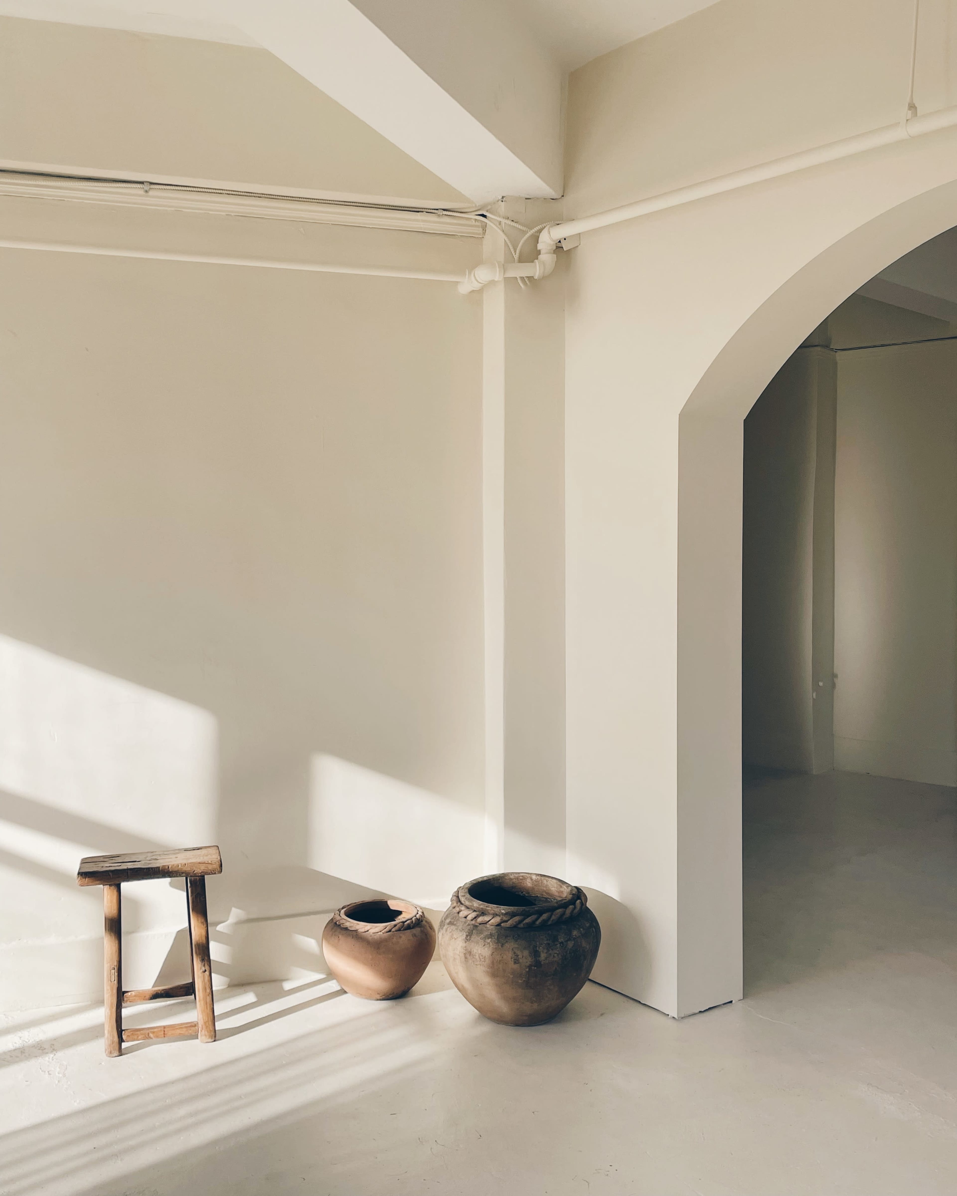 The image shows a minimalist room with a wooden stool and two clay pots positioned against a light-colored wall, illuminated by natural sunlight casting shadows.