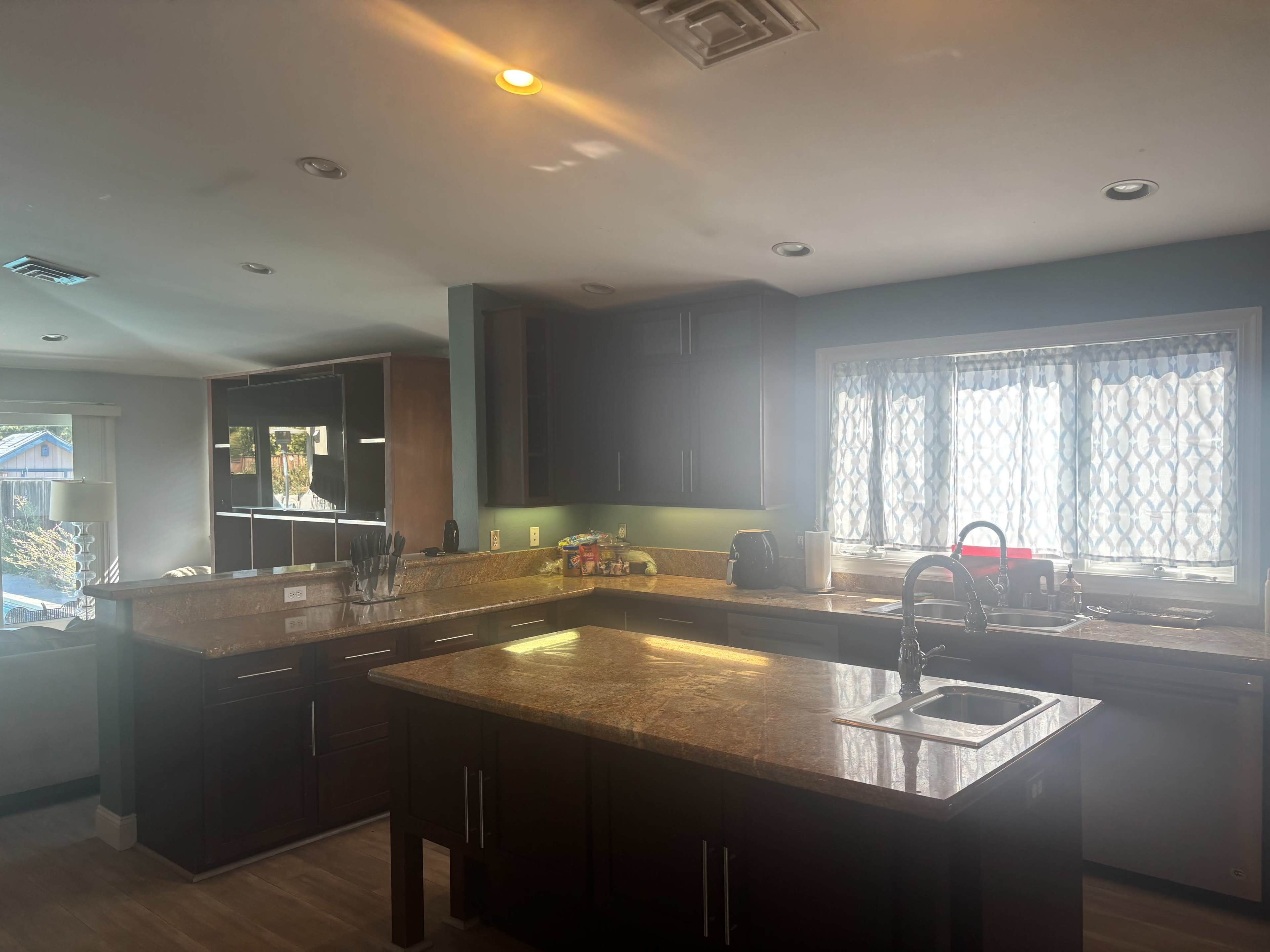 The image shows a modern kitchen featuring dark cabinetry, a central island with a sink, and a window with patterned curtains.