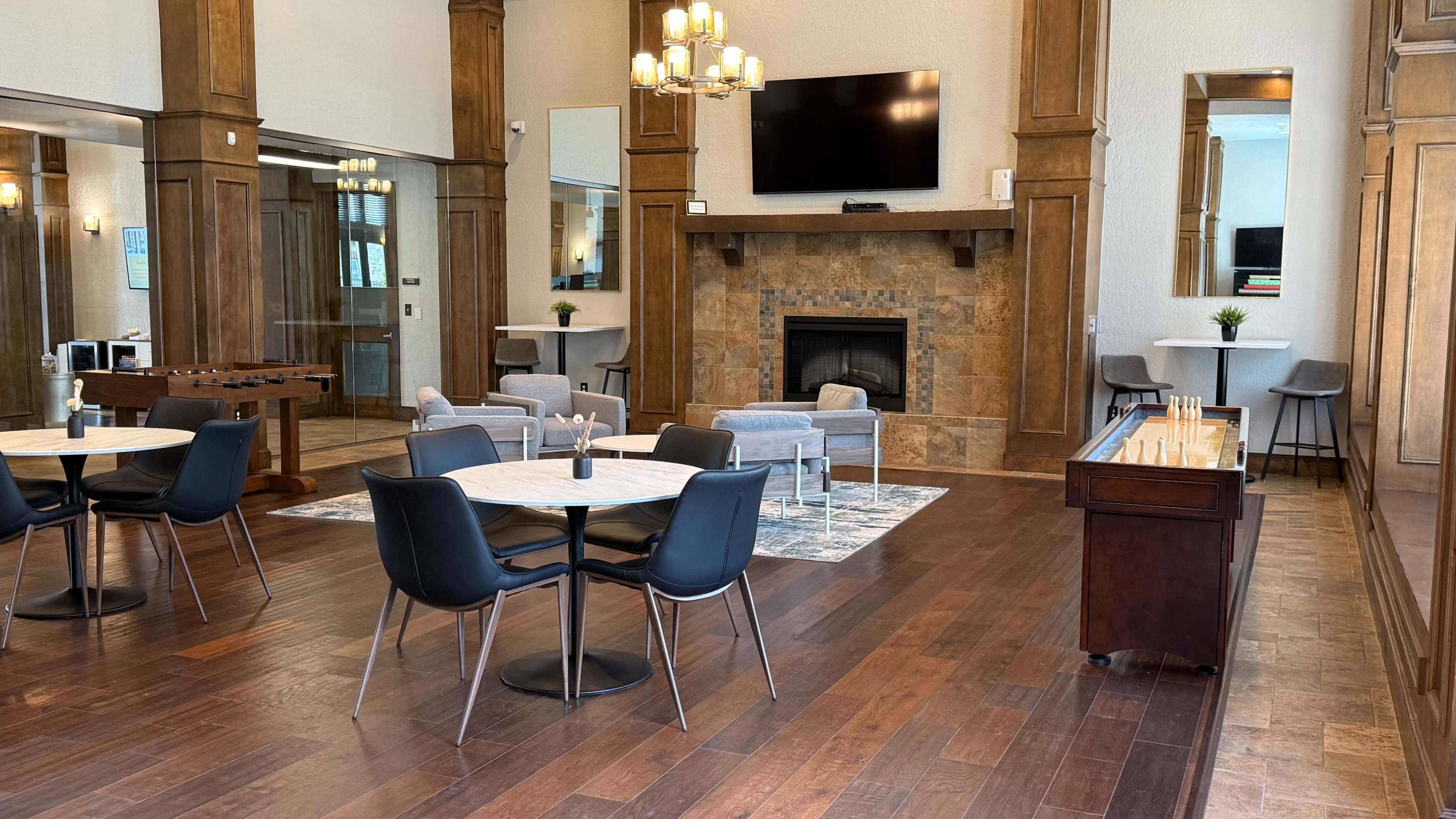 The image shows a spacious lounge area with wooden accents, featuring several seating arrangements, tables, a fireplace, and a large wall-mounted TV.