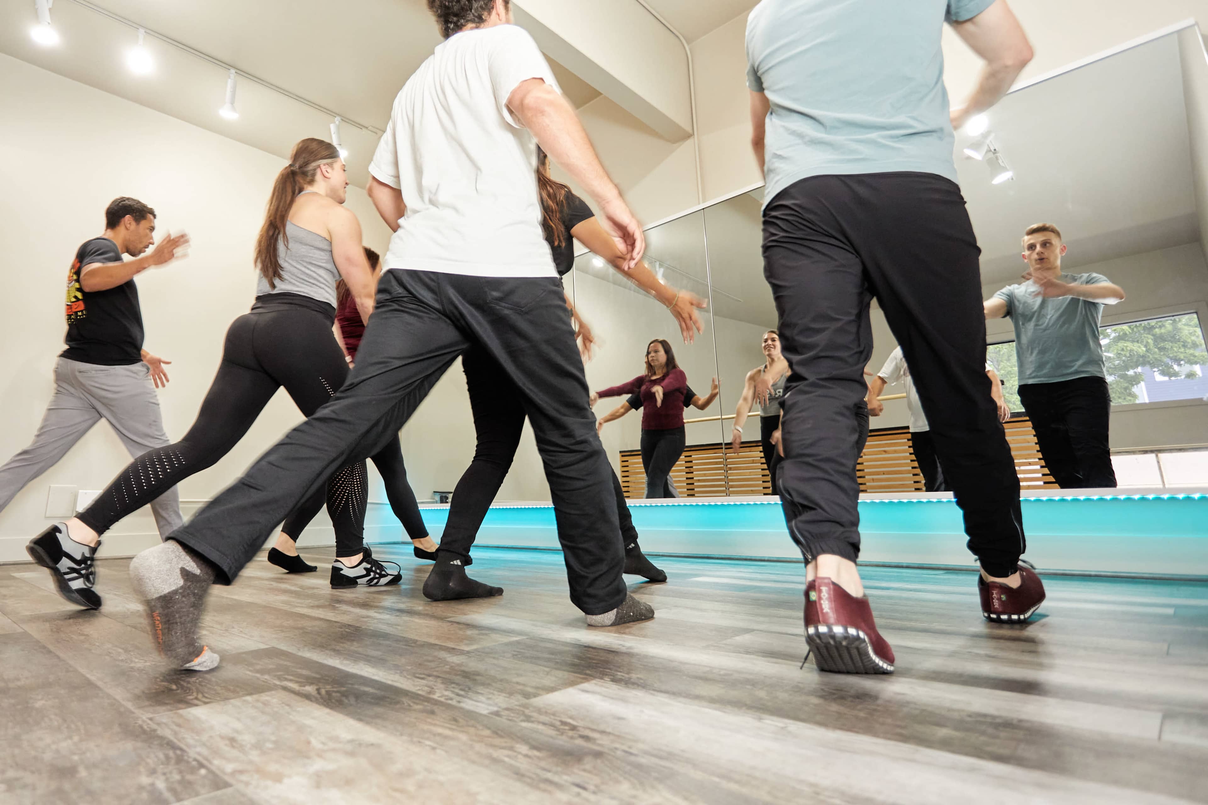 A group of dancers practice their movements in a well-lit studio with mirrors and a wooden floor.