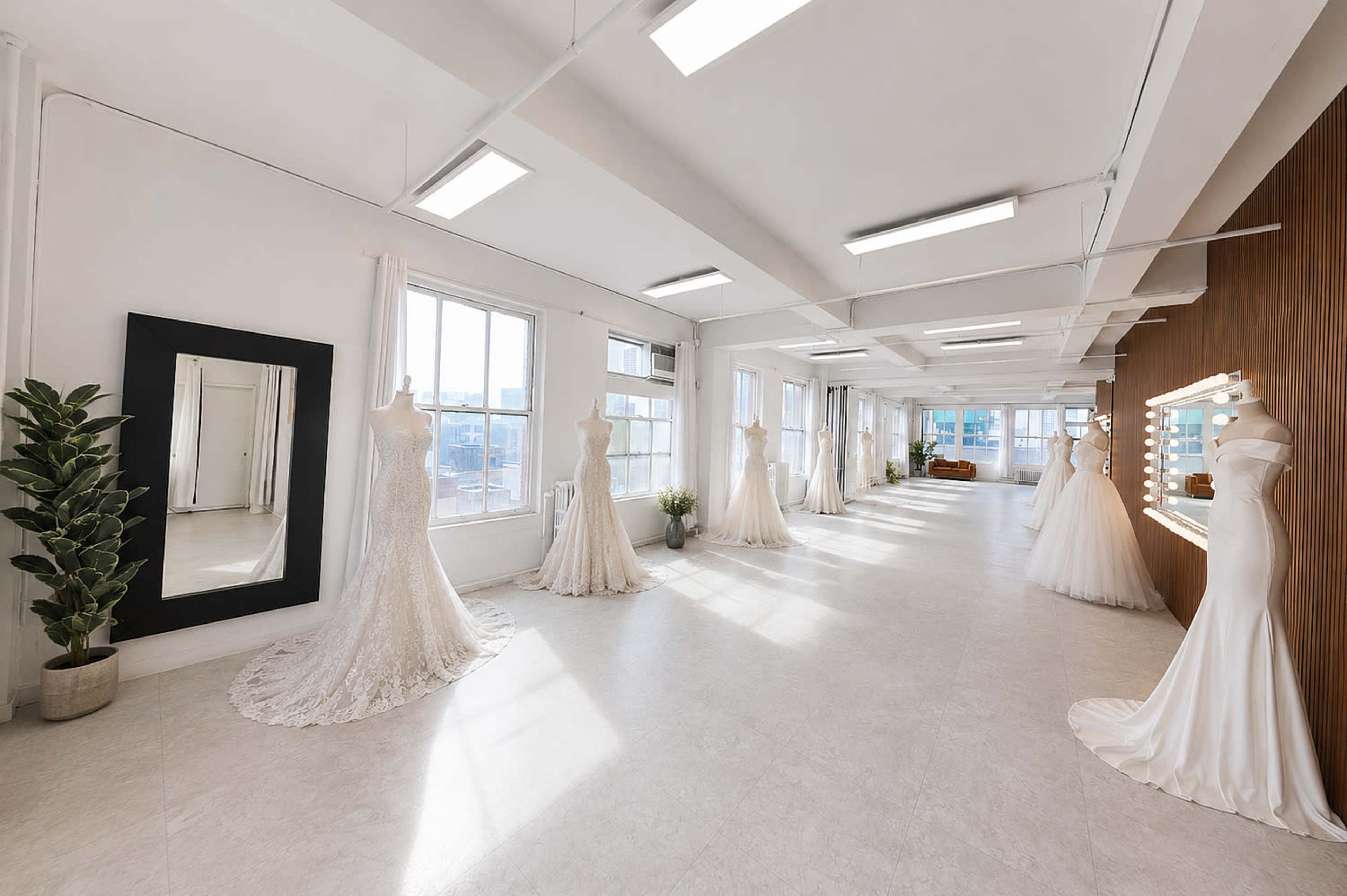 The image depicts a spacious bridal studio with several elegant wedding dresses displayed on mannequins, surrounded by large windows and minimalist decor.