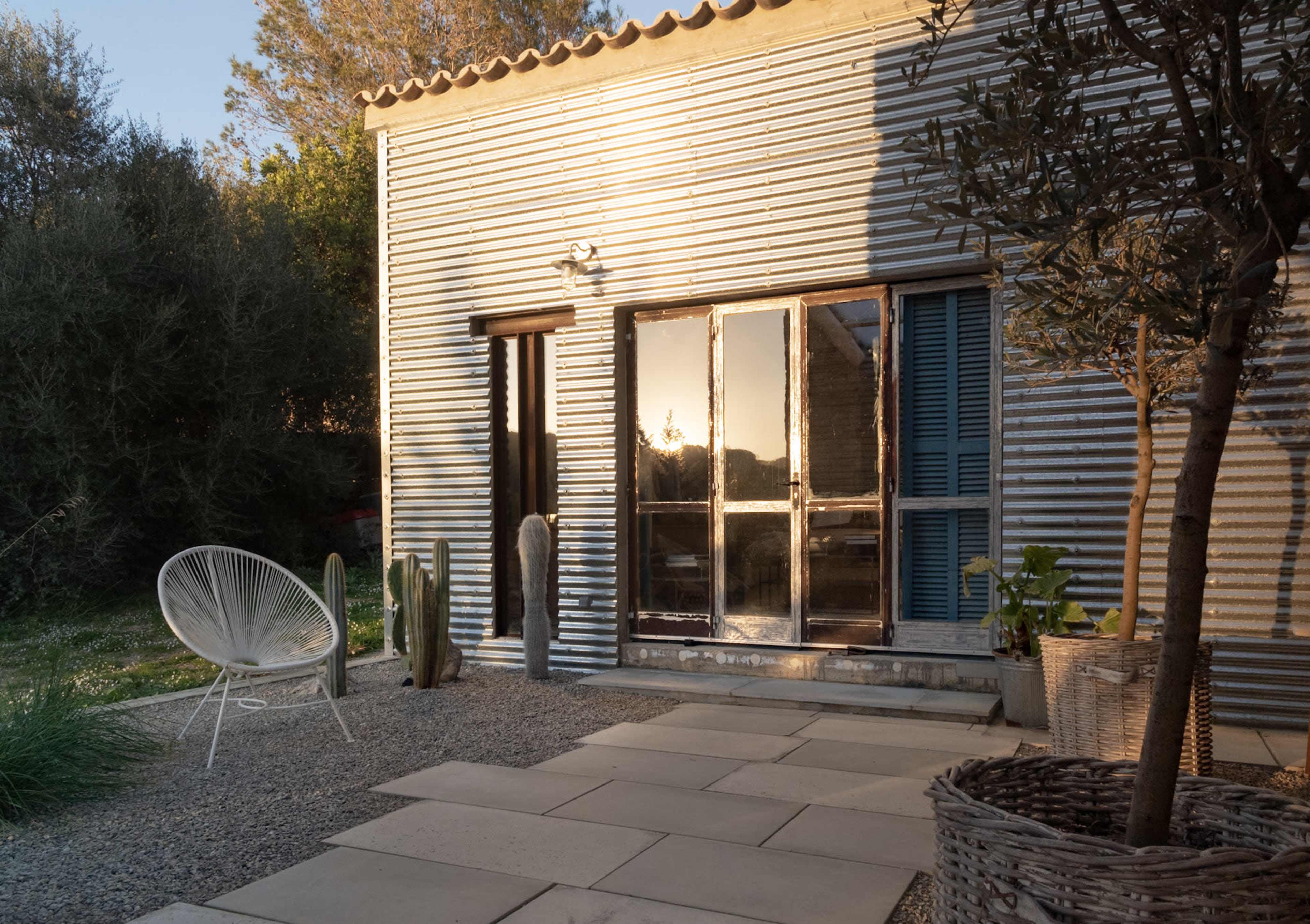 The image shows a corrugated metal house exterior with a patio area featuring stone tiles, a white chair, cacti, and potted plants.