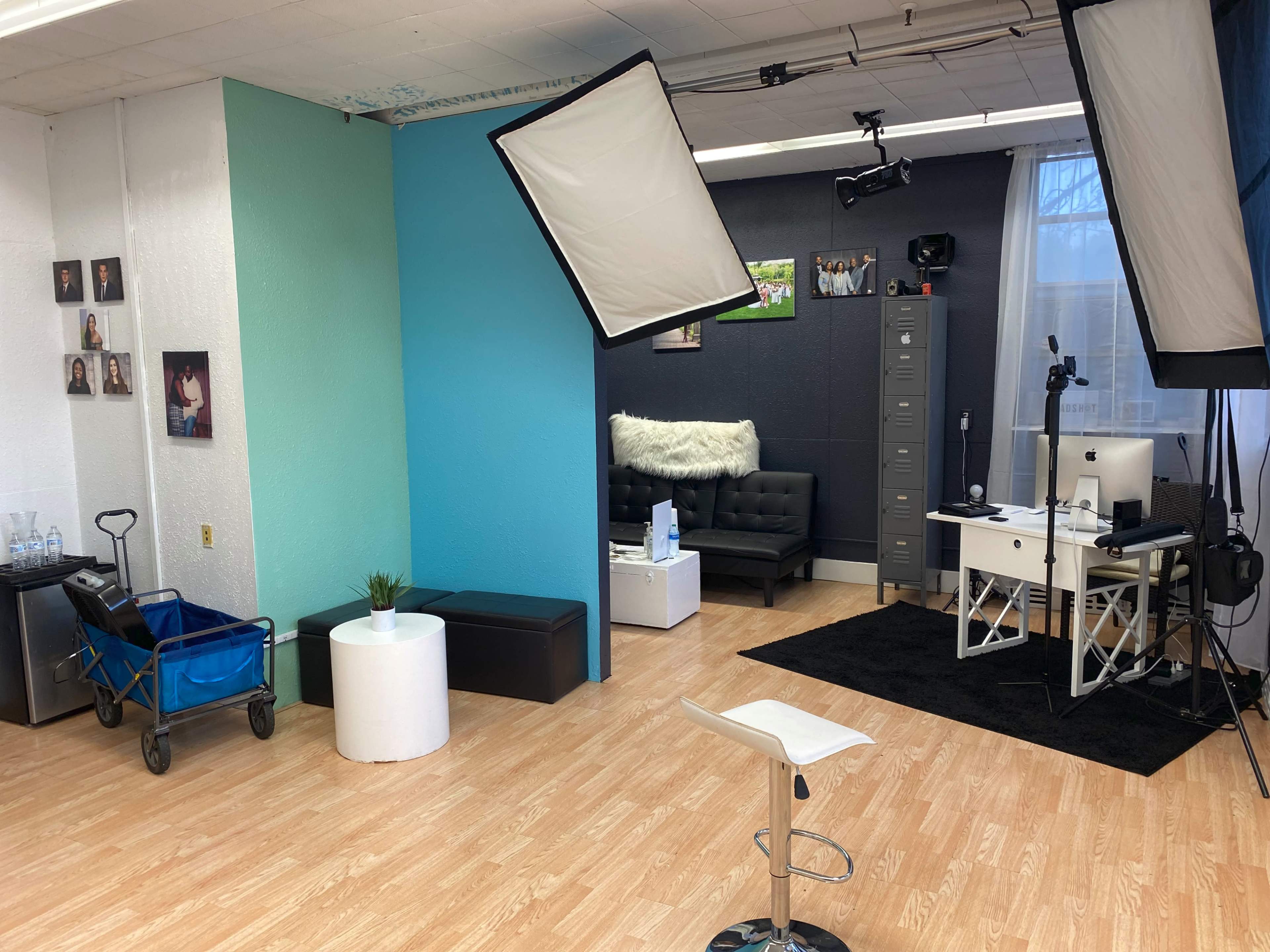 A spacious studio with different colored wall sections, a black couch, a white desk with a computer, and lighting equipment set up for a photoshoot.