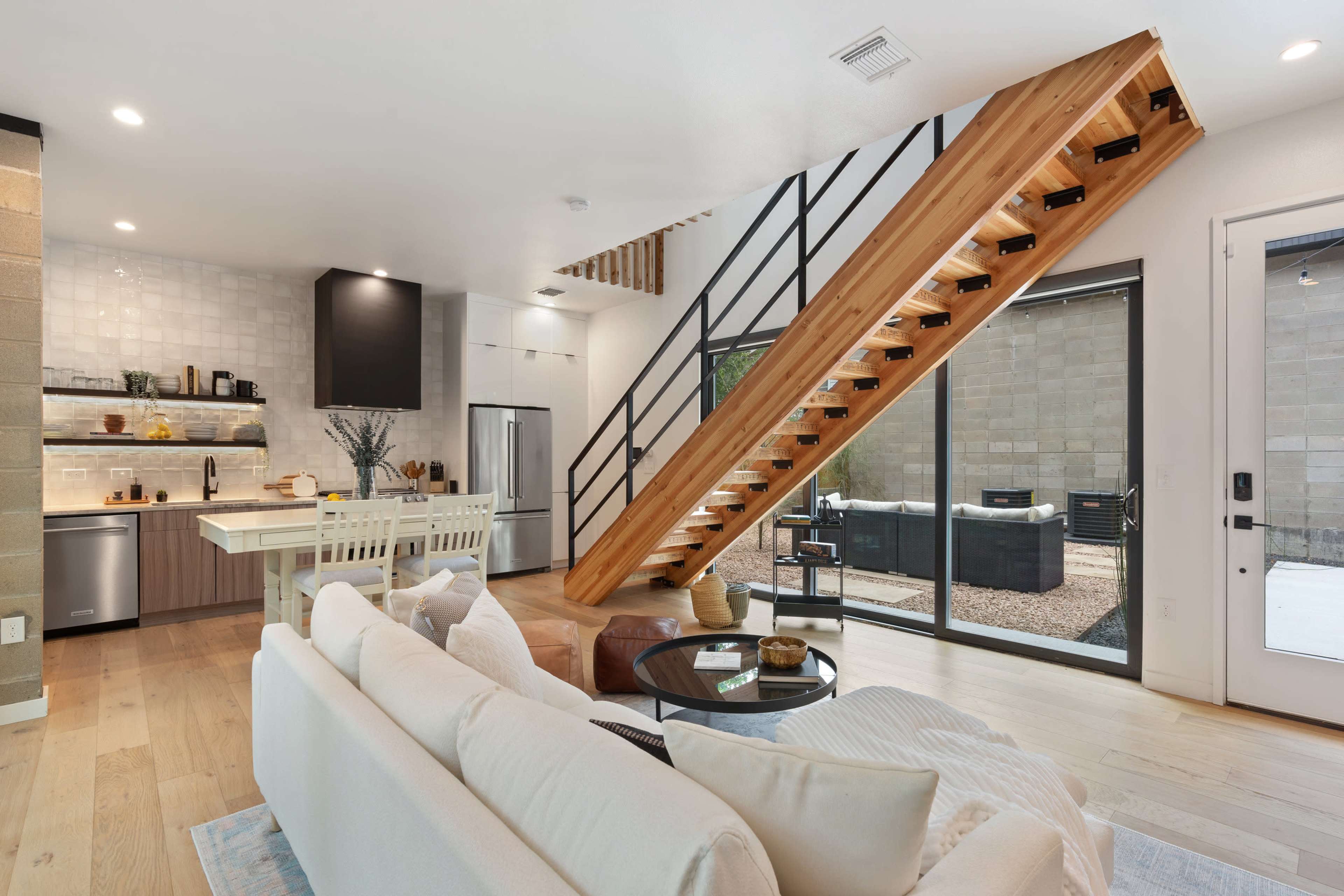 A modern interior features a living area with a white sofa, a kitchen with stainless steel appliances, and a wooden staircase leading to a second floor.