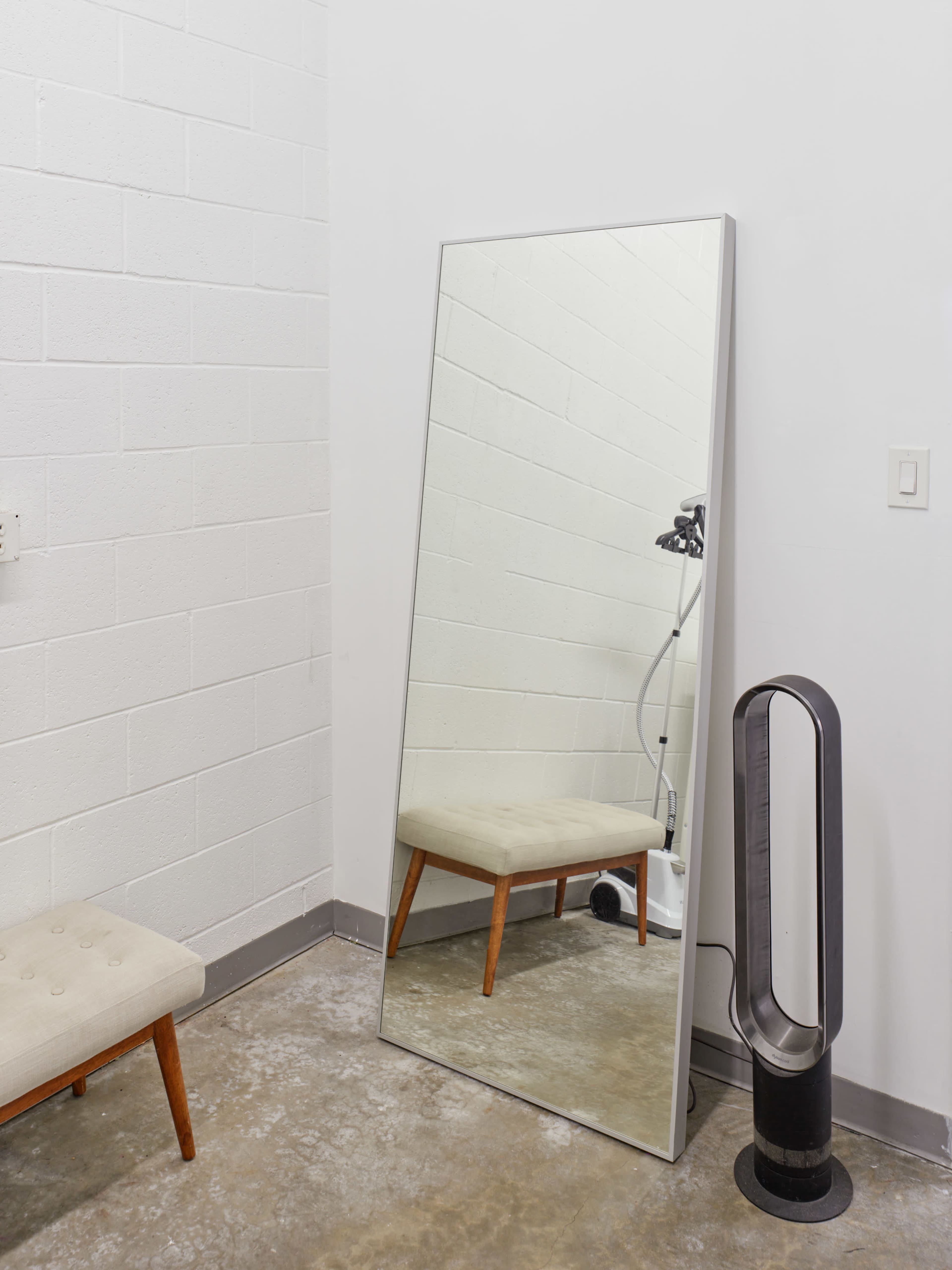 A full-length mirror stands against a white wall beside a small upholstered bench and a sleek fan.