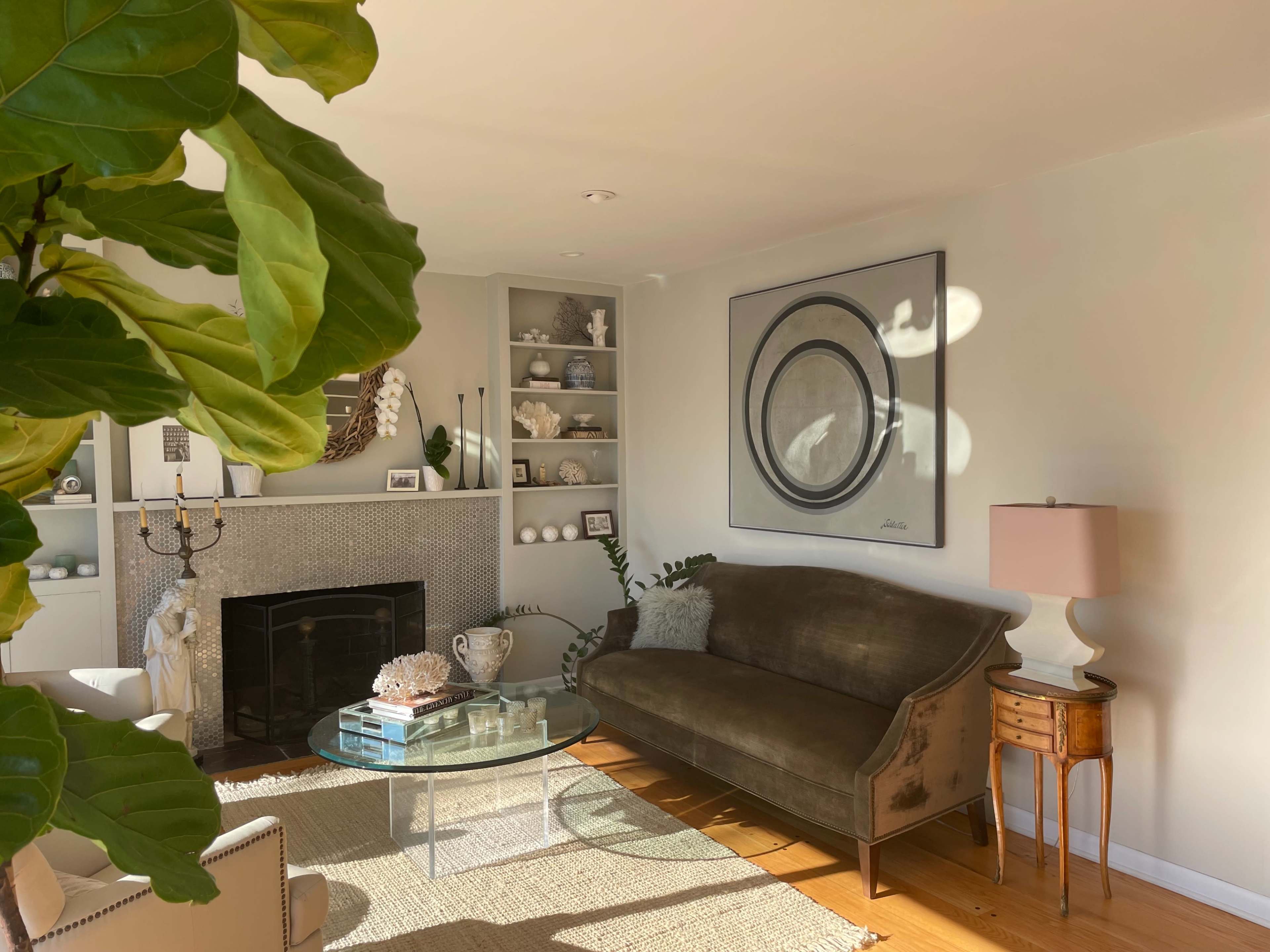 A cozy living room features a green velvet sofa, a round glass coffee table, and a decorative fireplace surrounded by shelving.