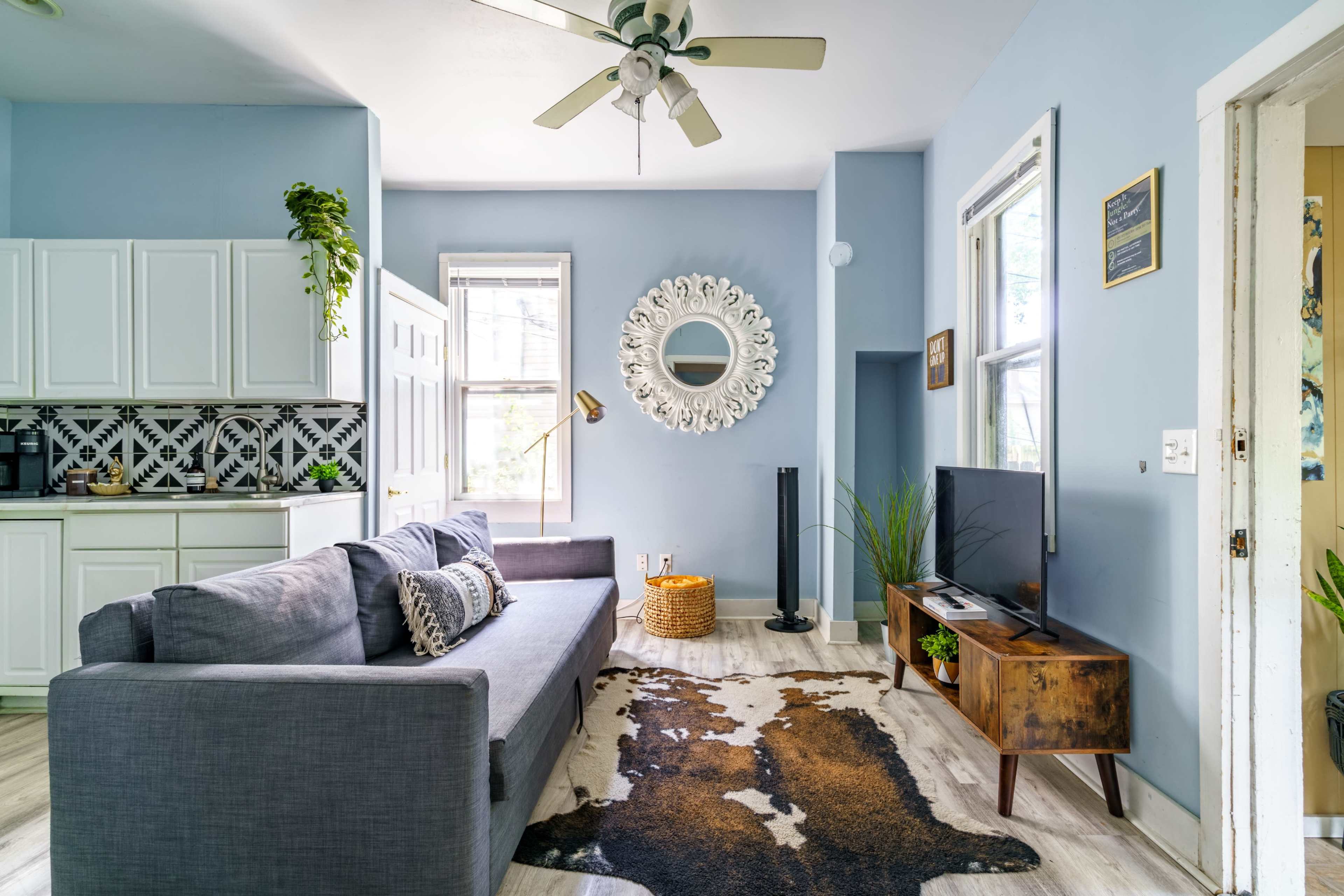 The room features a gray sofa facing a television, with a kitchen area in the background and a decorative mirror on the wall.