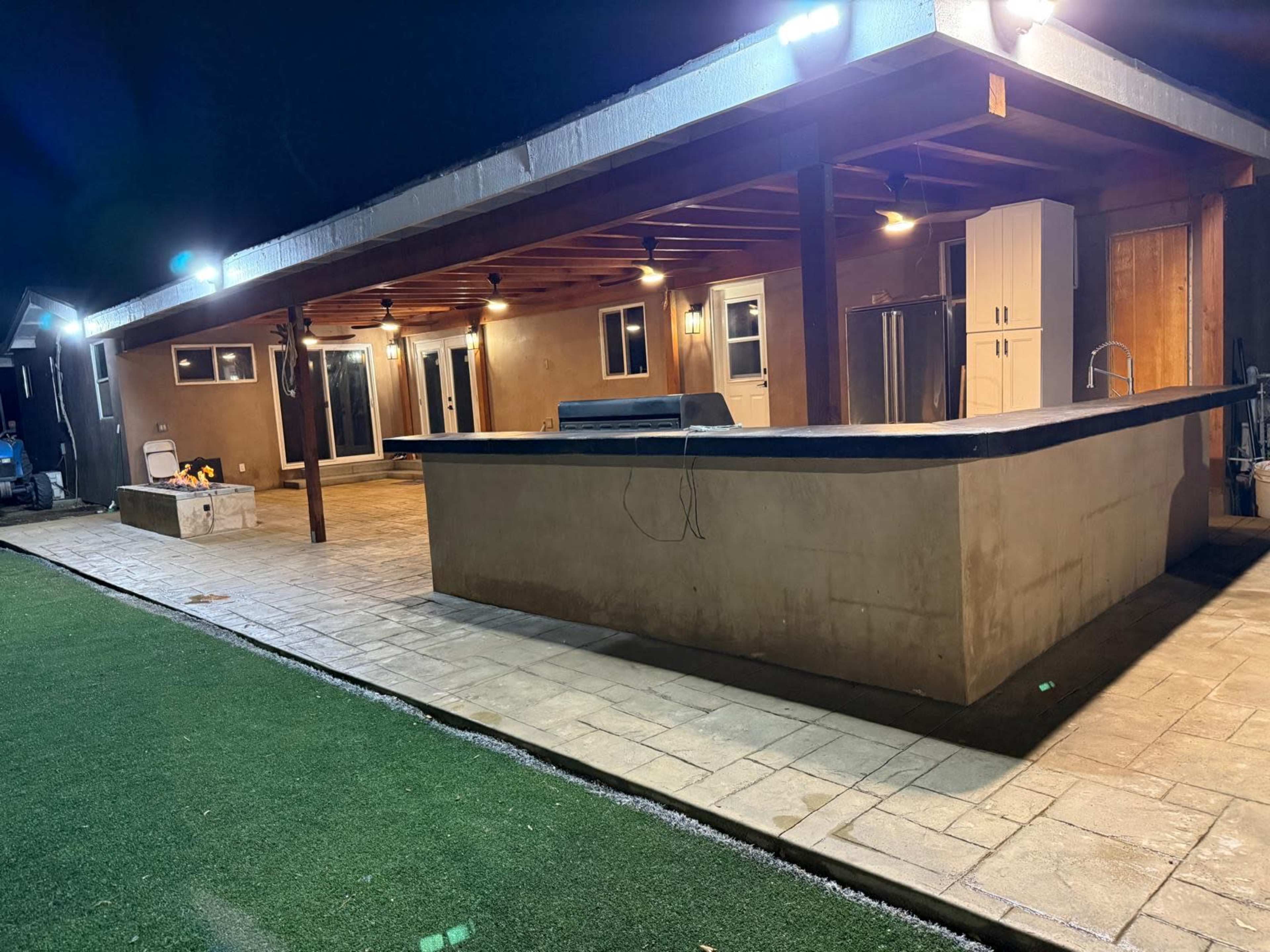 The image shows a modern outdoor kitchen and patio area illuminated at night, featuring a concrete countertop, built-in appliances, and a spacious paved area.