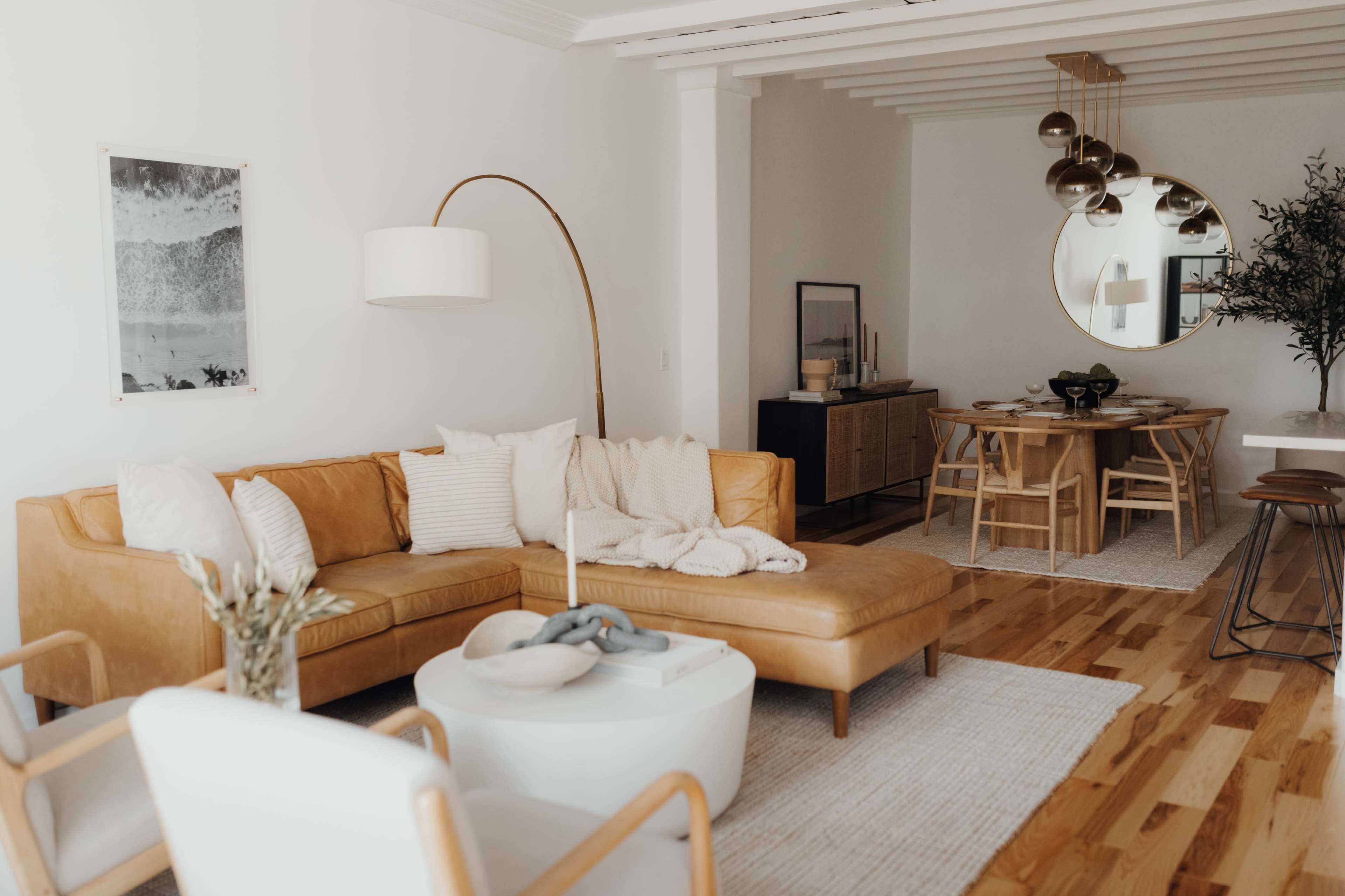 A modern living room with a brown leather sectional sofa, a round white coffee table, and a dining area featuring a wooden table and chairs.