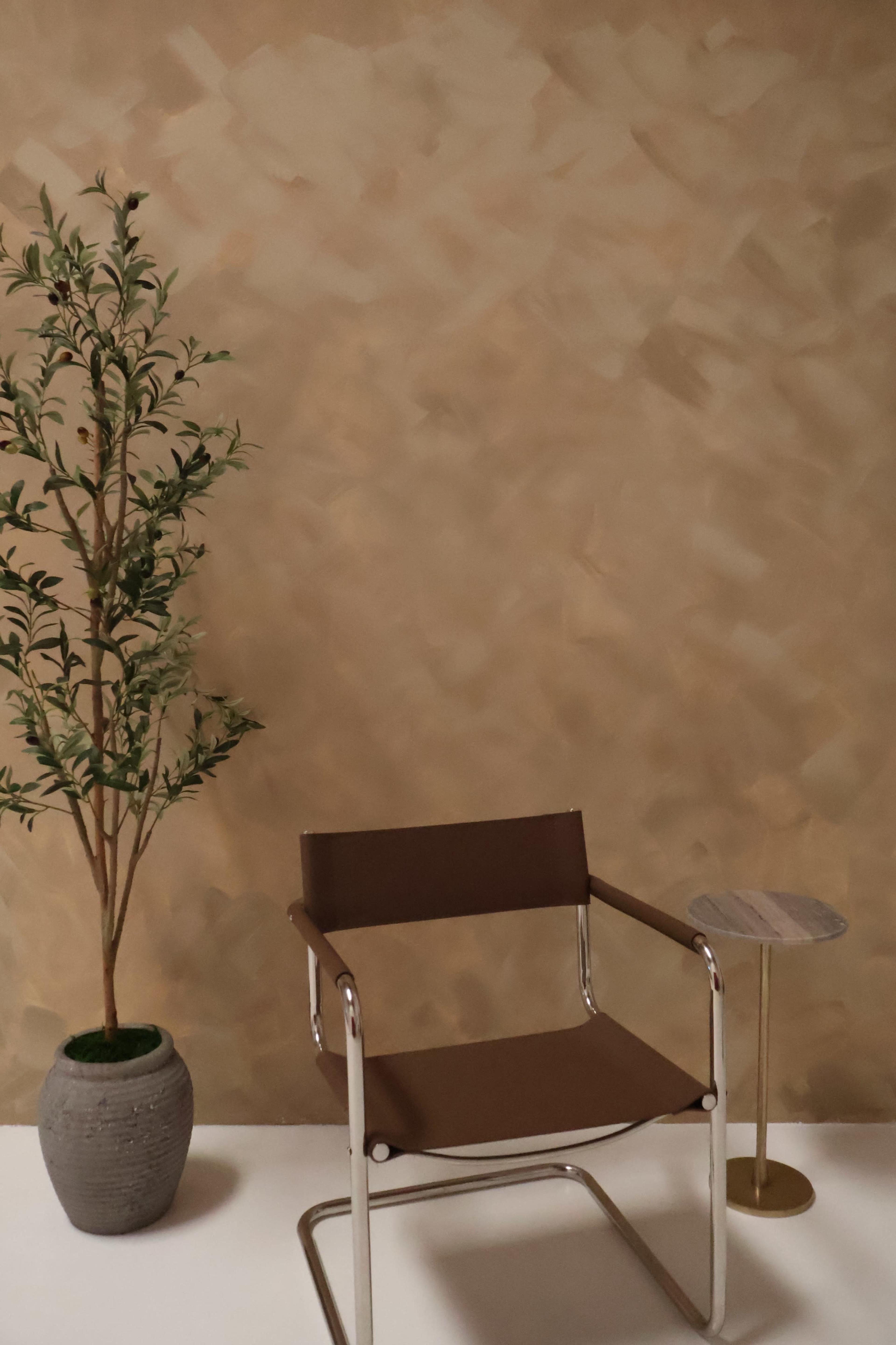 The image shows a modern chair beside a small side table and a potted plant against a textured beige wall.