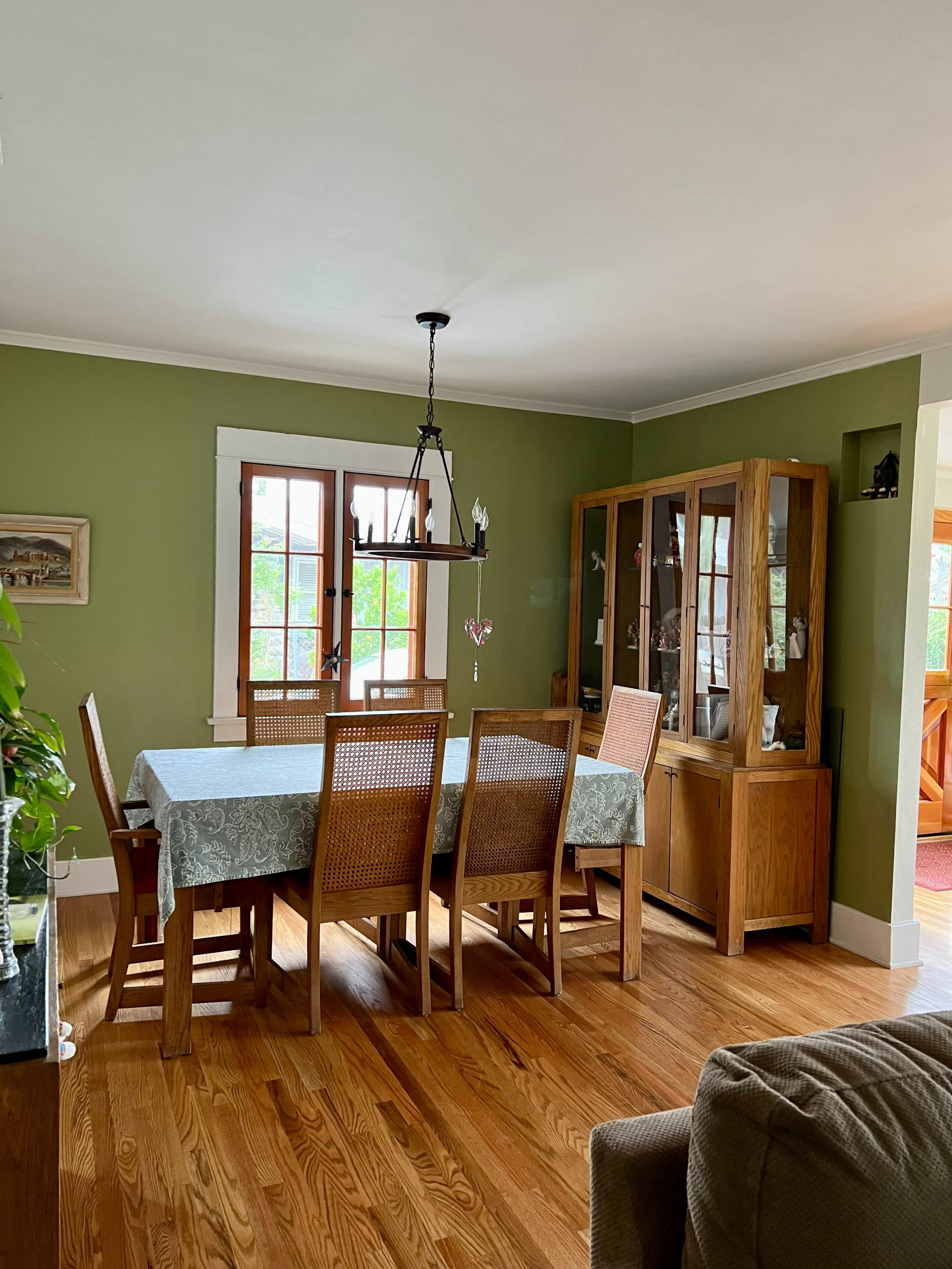A dining room features a large wooden table surrounded by chairs, a glass-fronted cabinet, and green walls with natural light streaming in through windows.