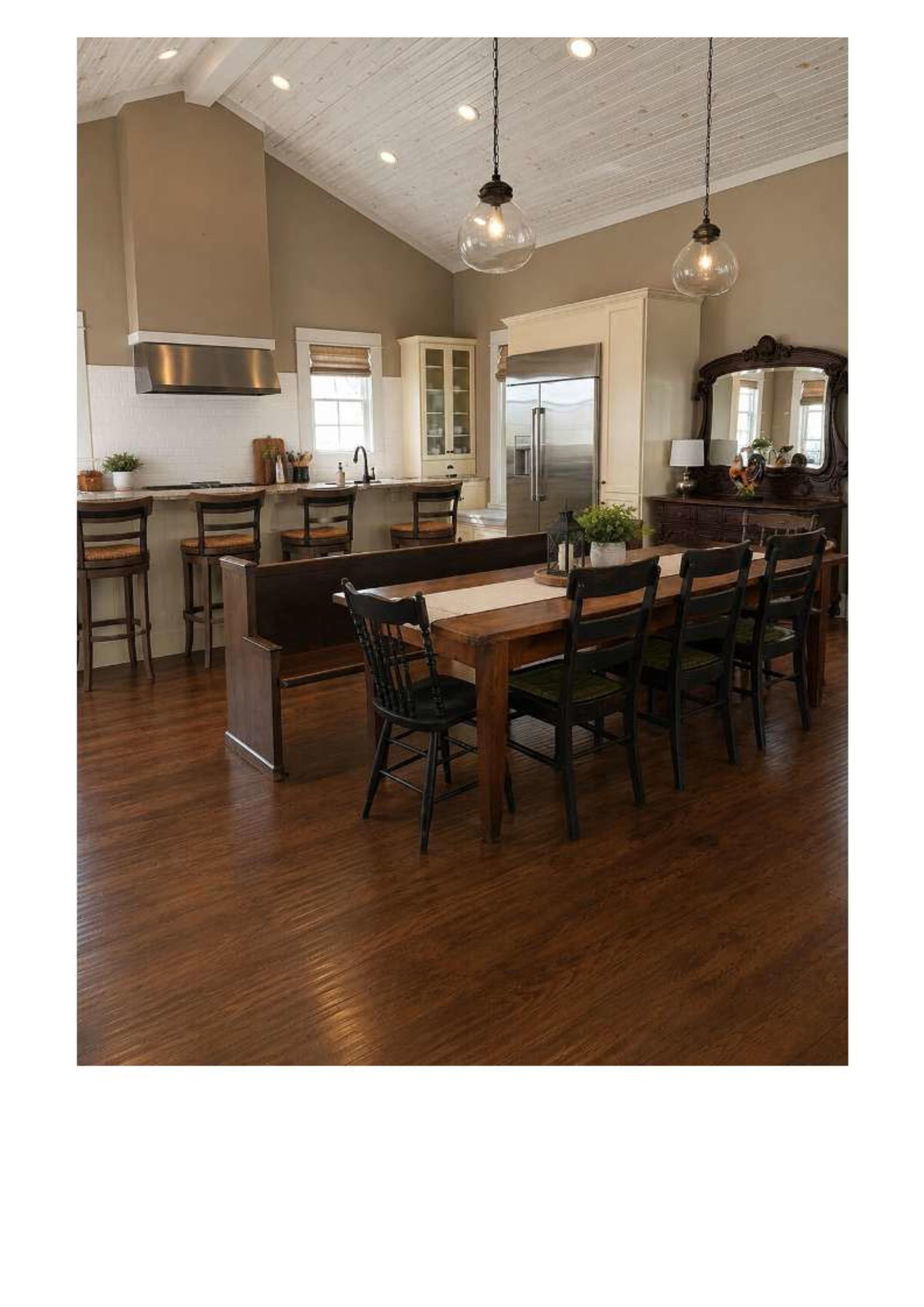The image shows a spacious kitchen and dining area featuring a long wooden dining table, black chairs, and a well-equipped kitchen with bar seating and modern appliances.