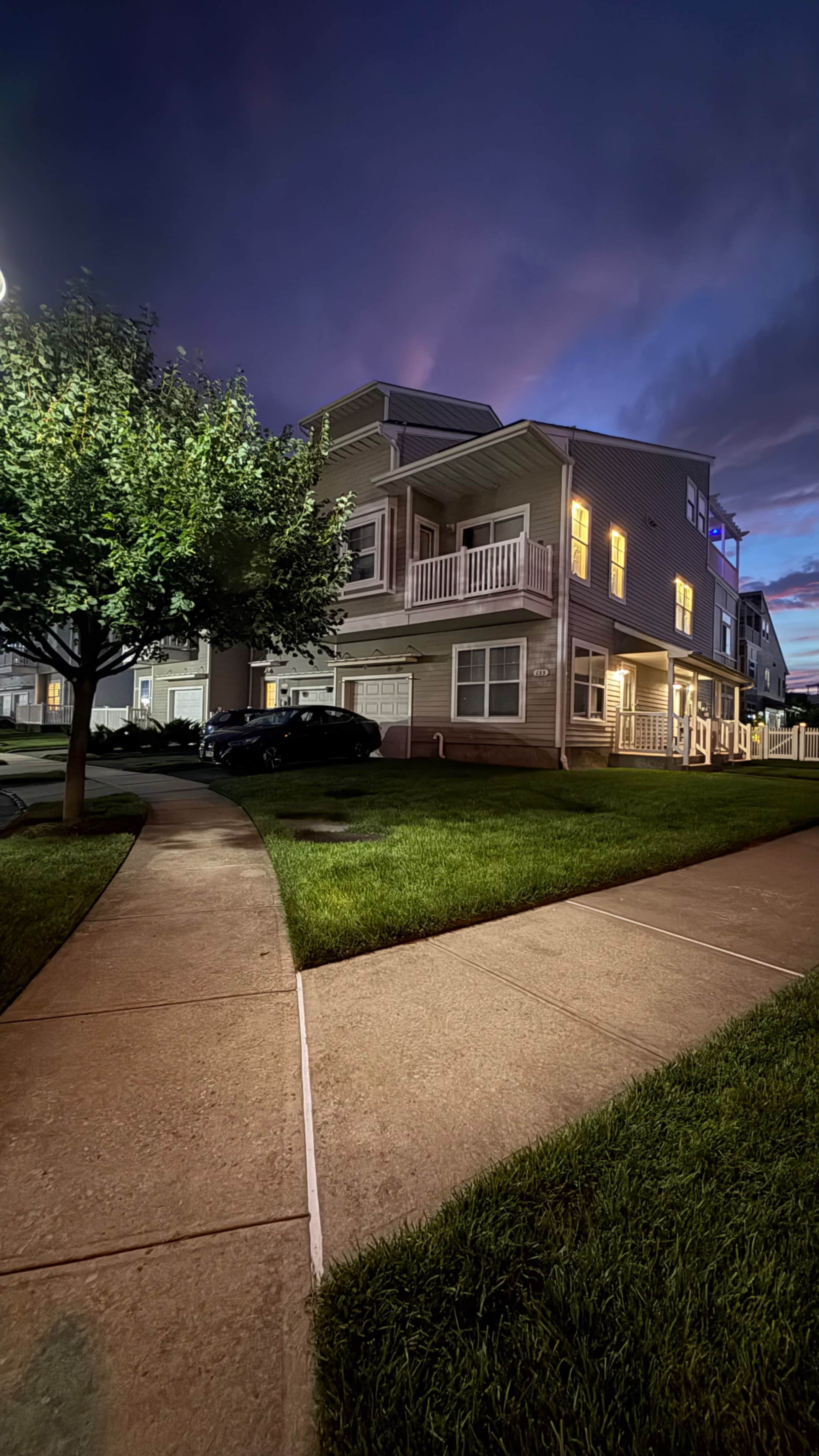 A two-story house with illuminated windows is set against a twilight sky, surrounded by a landscaped lawn and a sidewalk curving around the property.
